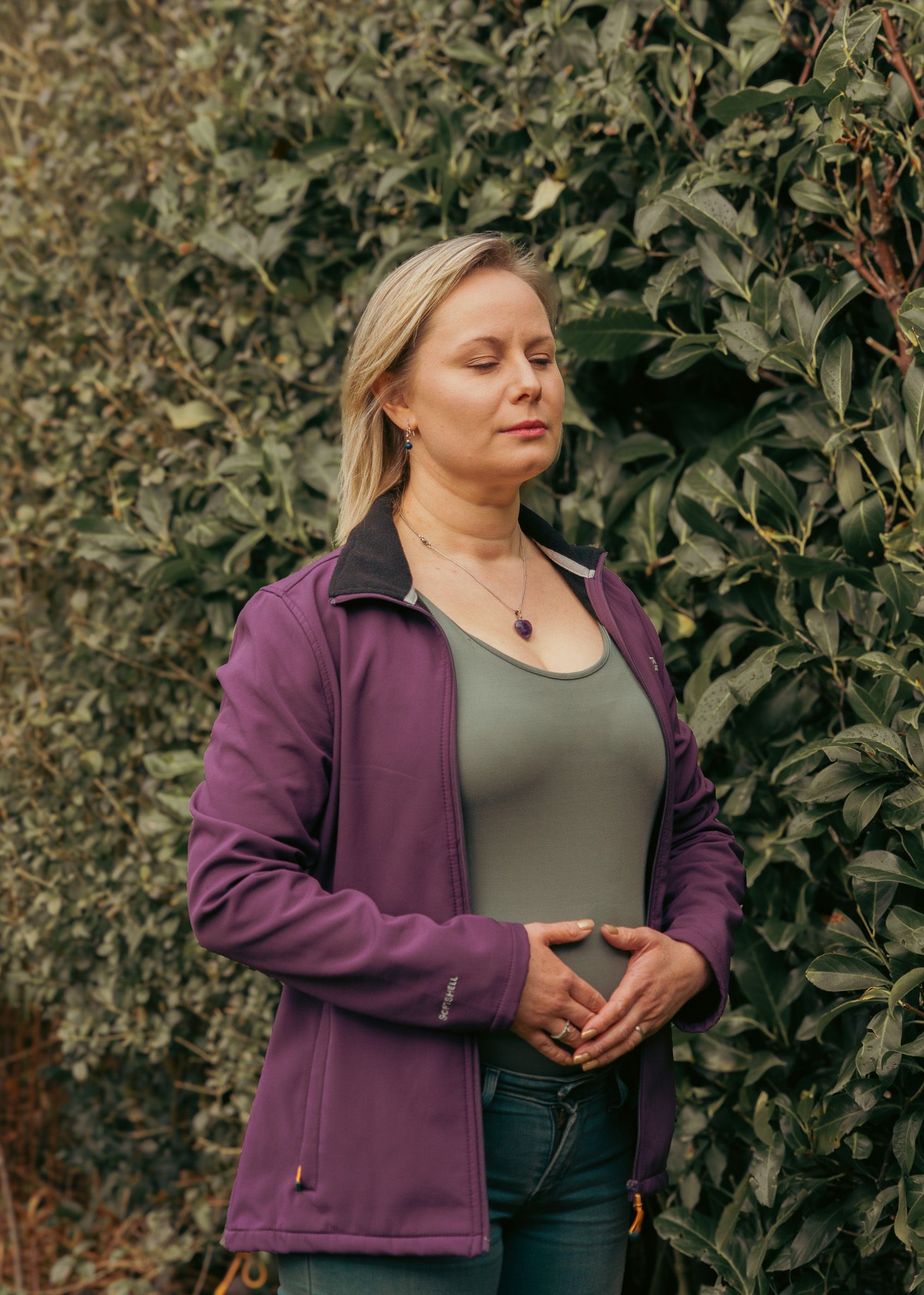 Person in purple jacket standing thoughtfully against green foliage backdrop.
