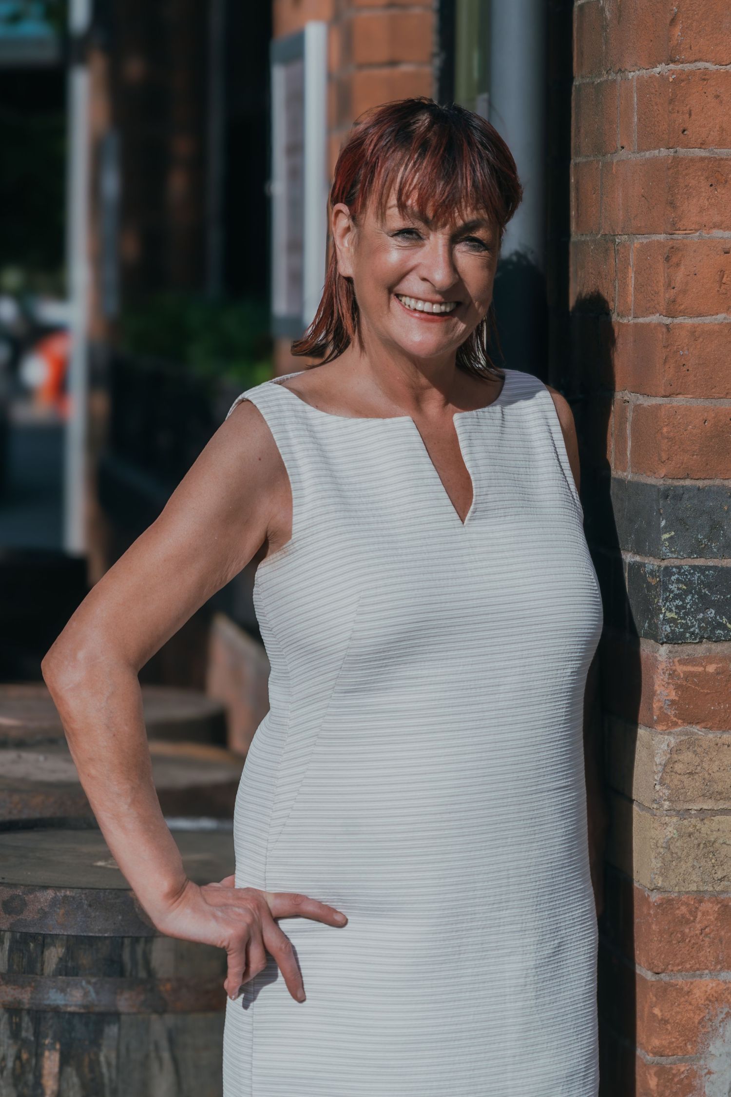 Personal Brand Photography. Business owner wearing a white sleeveless dress poses with a bright smile against a brick wall background.