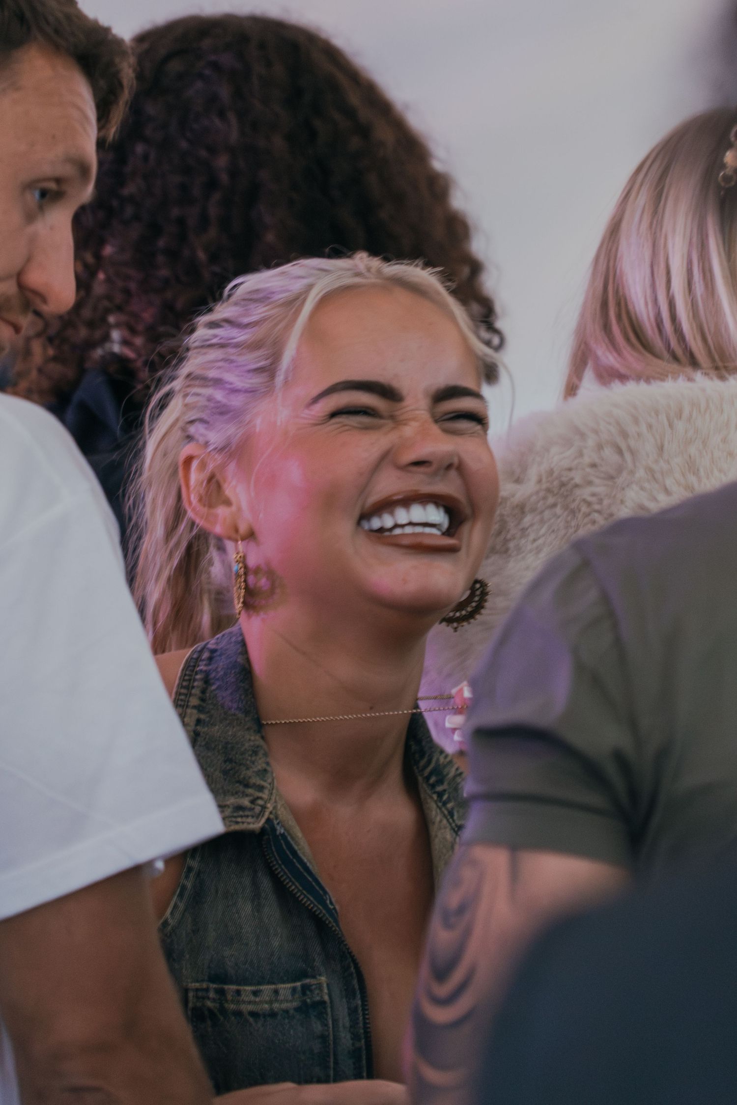 Young woman with dyed platinum hair and a denim vest laughs joyfully among friends at a casual gathering.