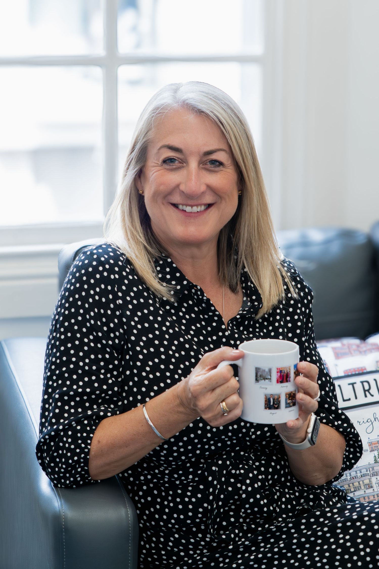 Woman in black and white polka dot blouse sits comfortably holding a coffee mug and smiling in bright room.