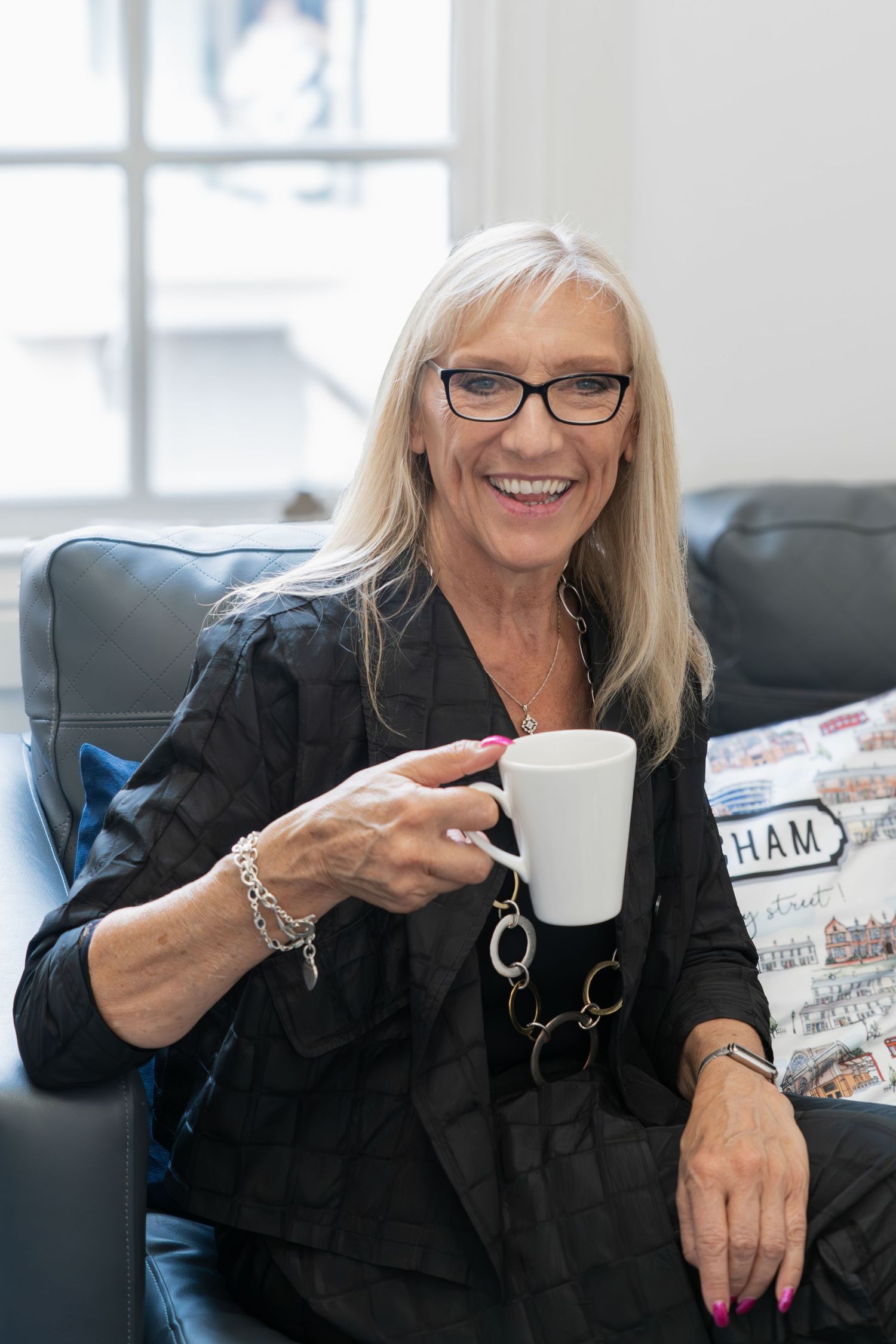 Professional wearing black attire and glasses sits in a chair holding a white coffee mug while smiling warmly.