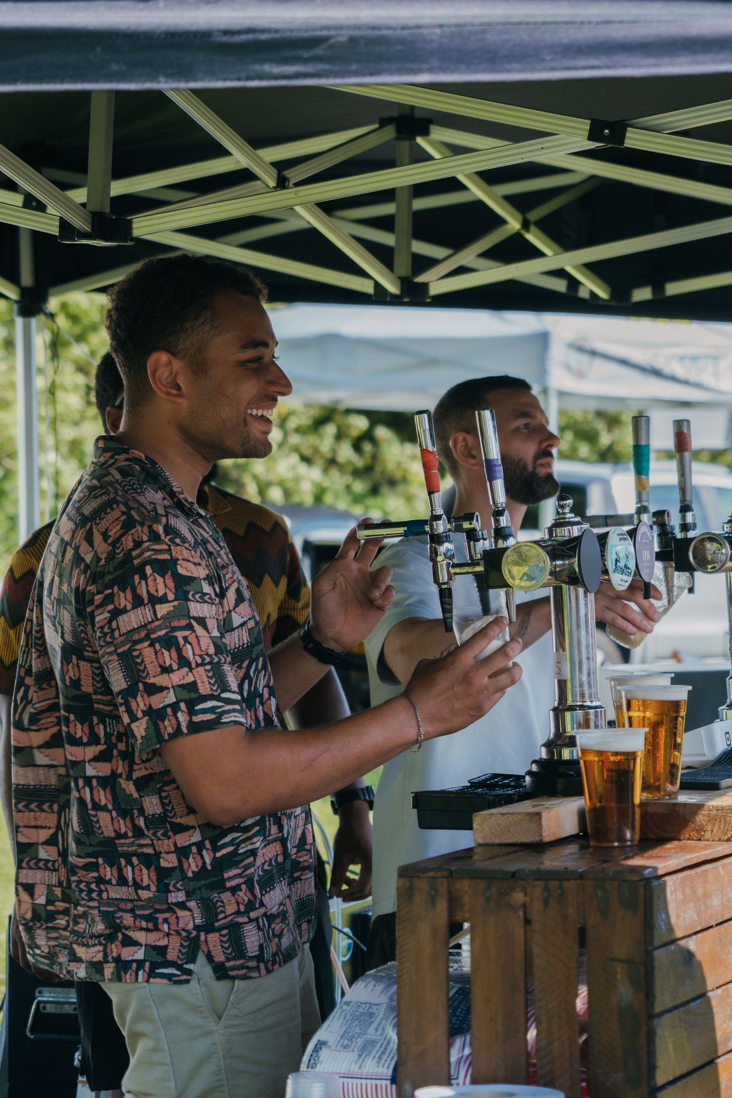 Bartenders pour draft beers from taps at an outdoor event under a pop-up tent canopy.