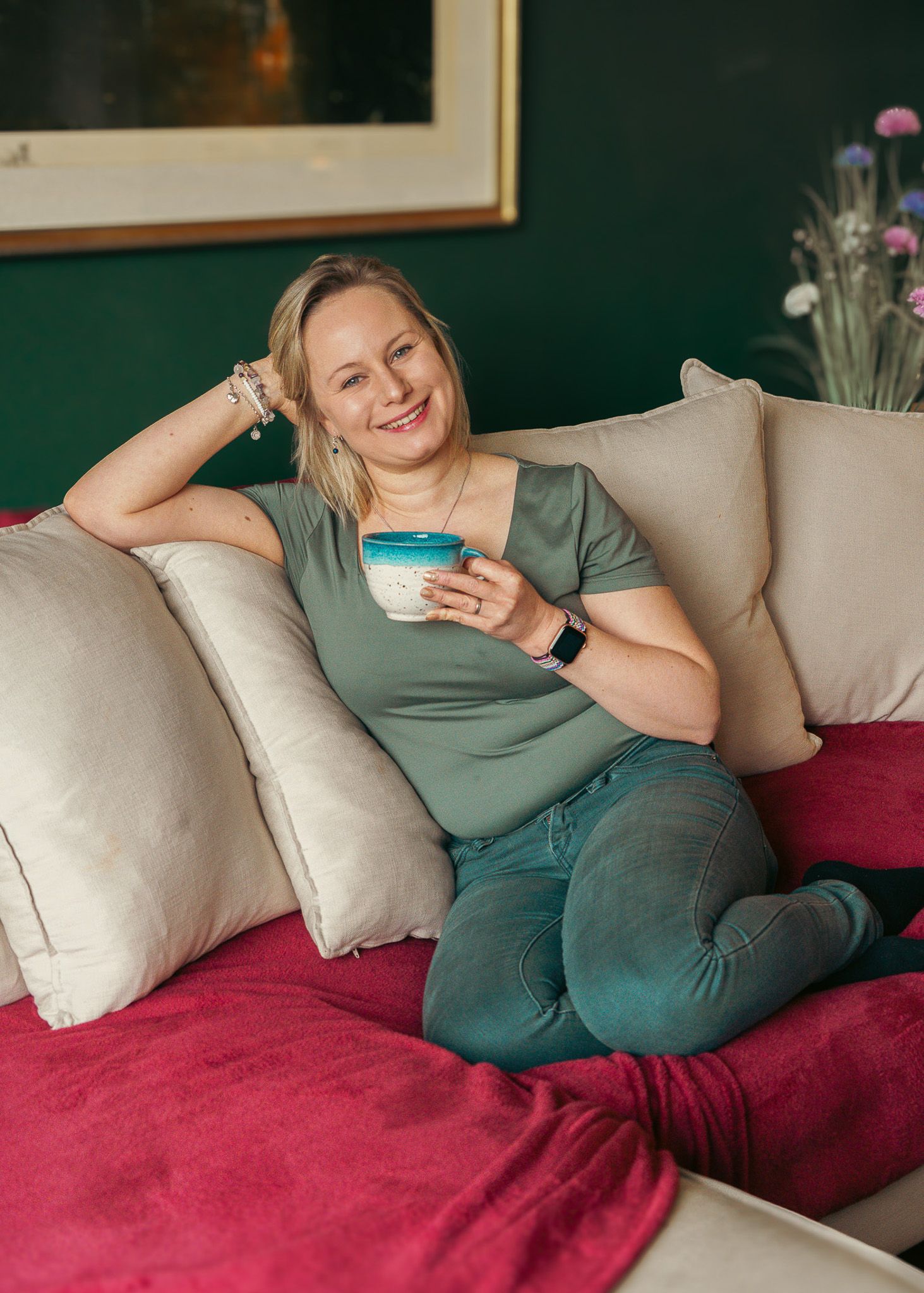 Person relaxing on a cozy couch with a red blanket while holding a blue cup.