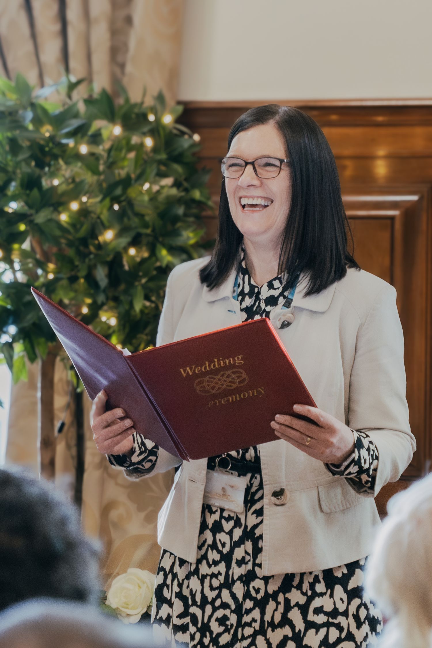 A wedding officiant wearing glasses and a beige blazer smiles while holding a red ceremony book in front of a Christmas tree.