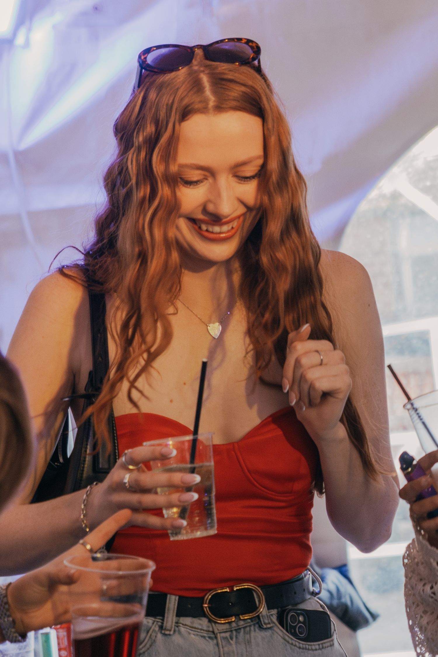 A woman in a red tank top laughs while holding a beverage at a social gathering with sunglasses on her head.