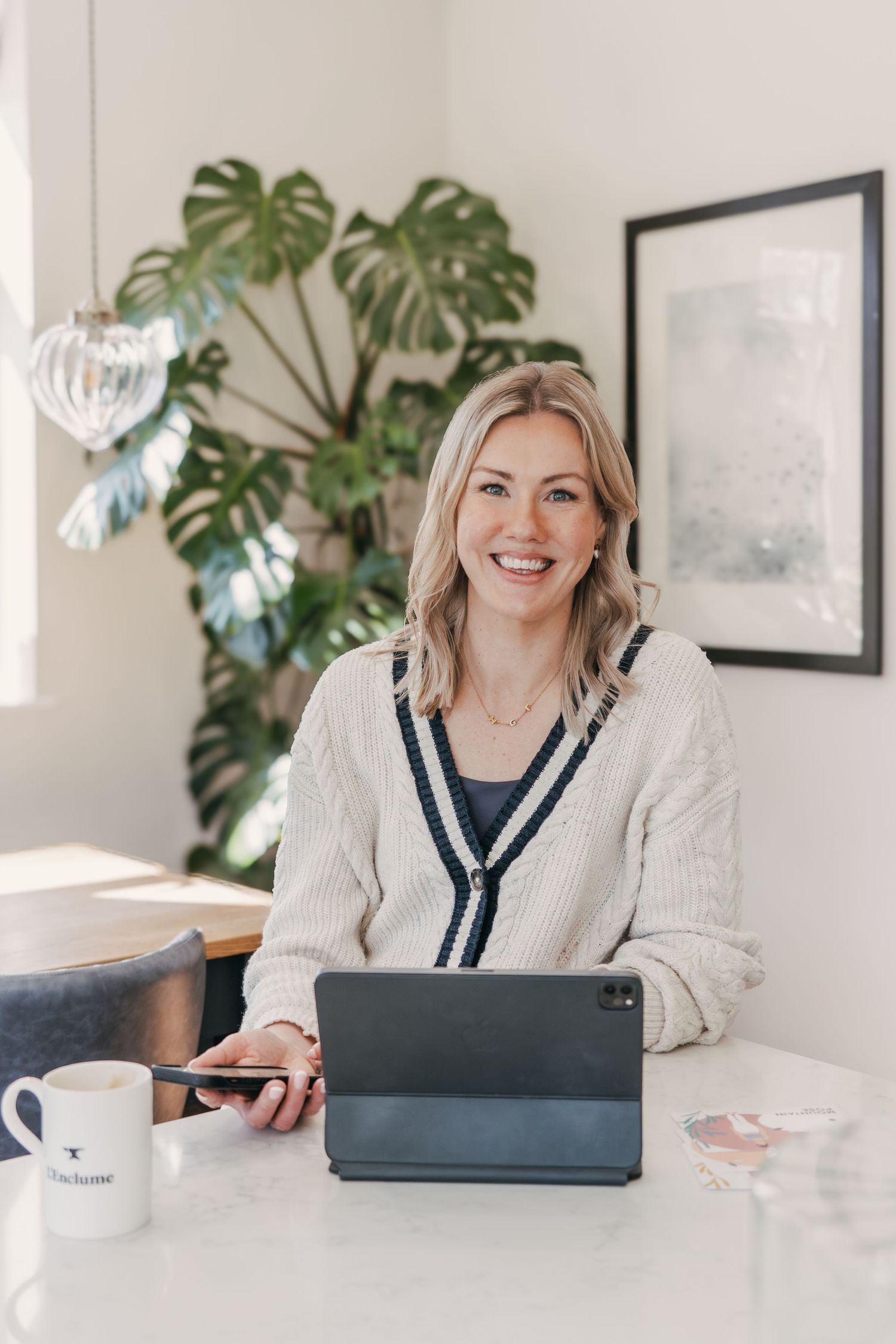 Person in preppy white sweater works on tablet device at bright desk with monstera plant and framed art in background.
