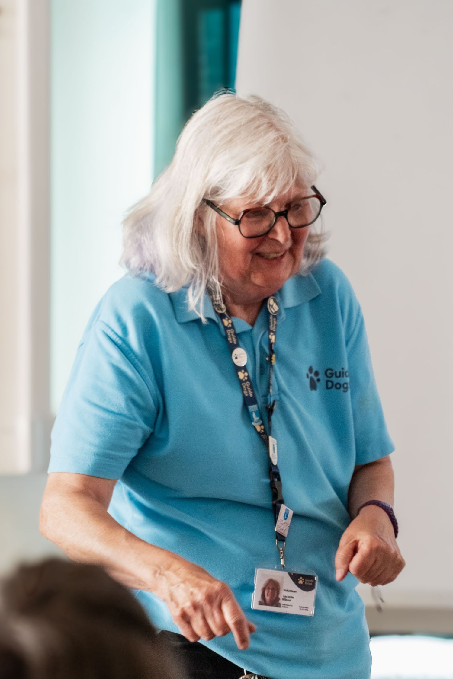 Healthcare worker in light blue uniform polo shirt and lanyard smiles while gesturing during conversation.