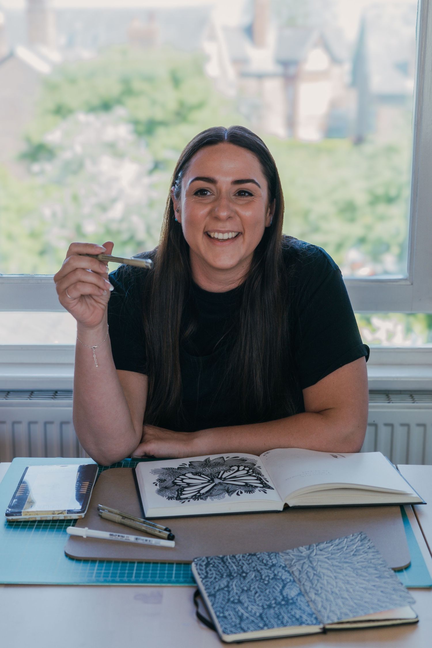 Personal Branding Photography for an artist who sits at desk with sketchbooks and art supplies while working on illustrations by window.