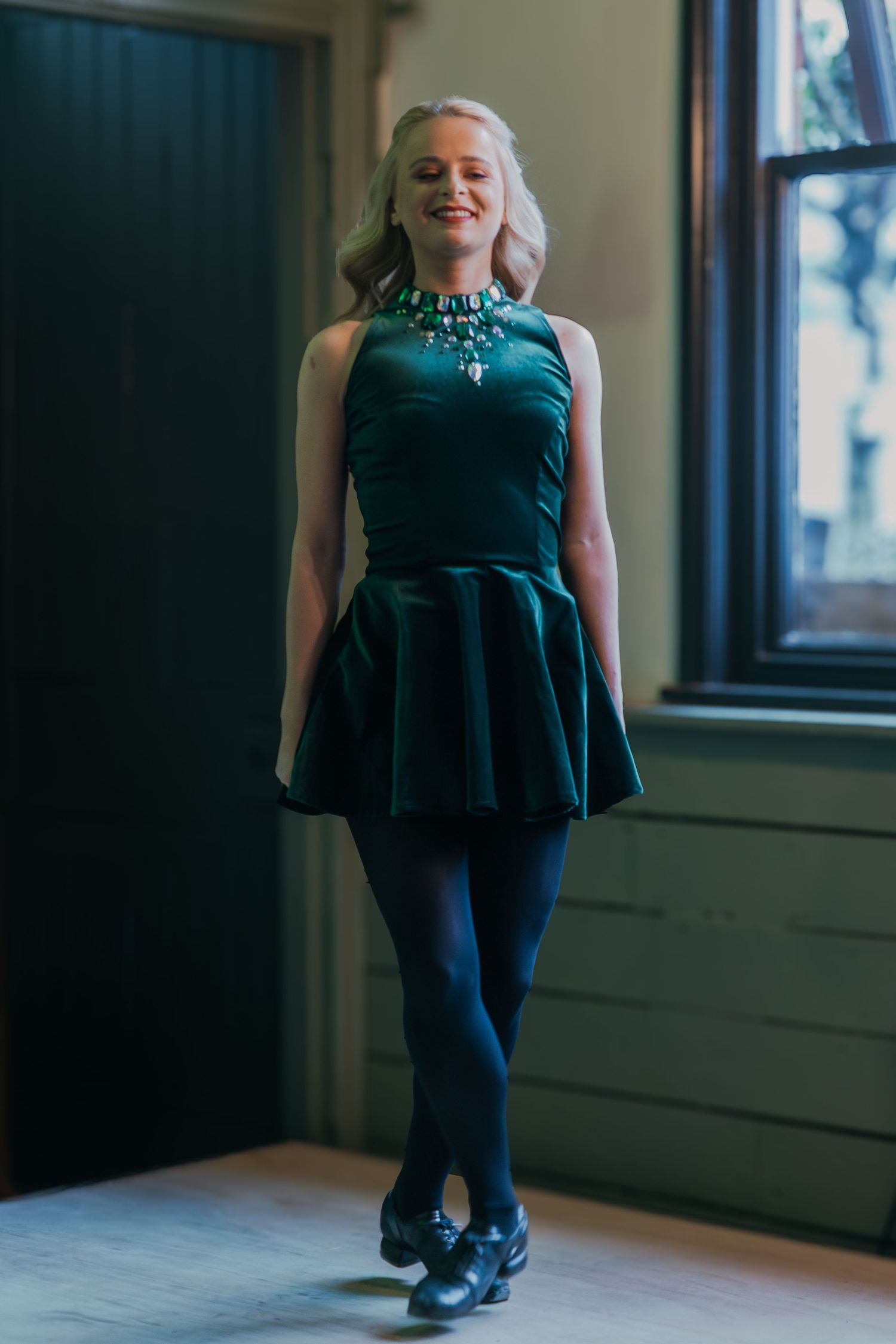 A person wearing a dark teal dress with pleated skirt, black tights and statement necklace stands by a window.