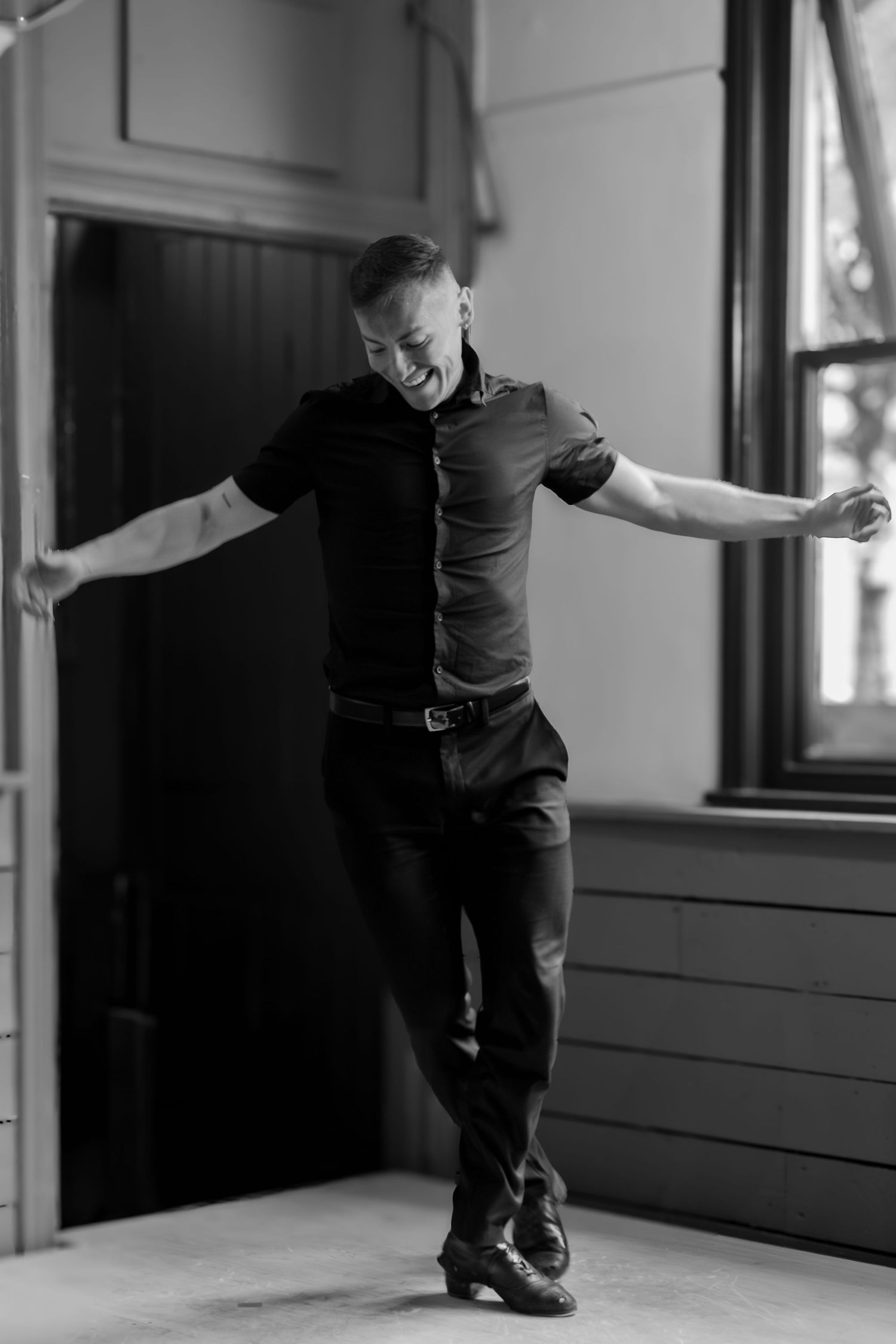 Black and white photo of a person dancing joyfully in a room with dramatic window lighting.