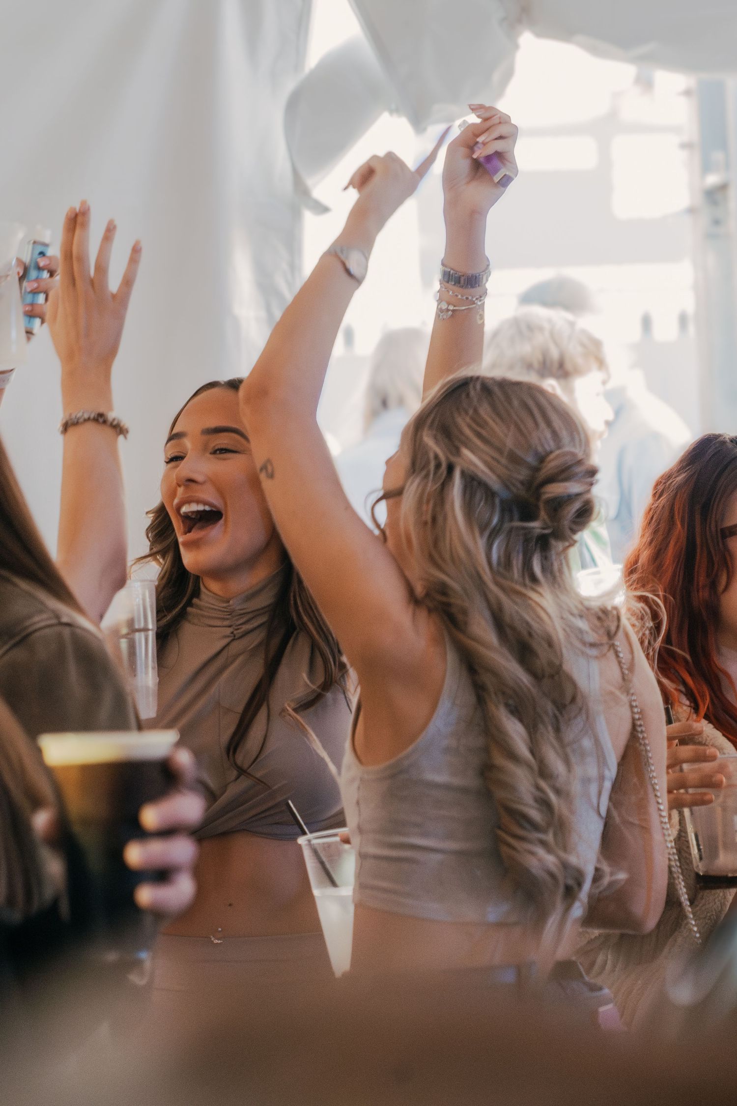 Group of friends dancing and celebrating with drinks at a lively party under white tent lighting.