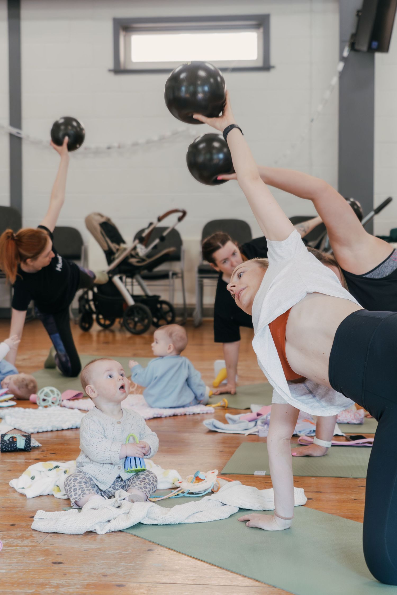 Fitness class for mothers doing kettlebell workouts while their babies play on mats in studio gym.