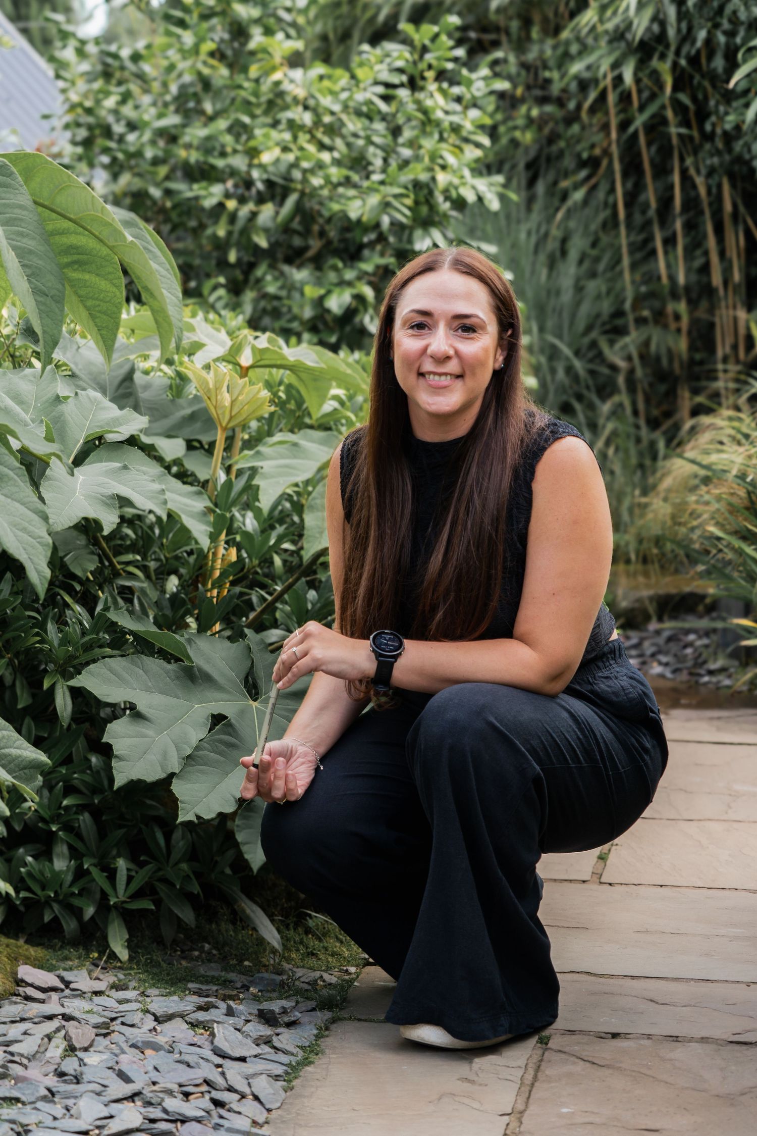 Someone in a black dress poses outdoors near lush green foliage and stone landscaping elements.