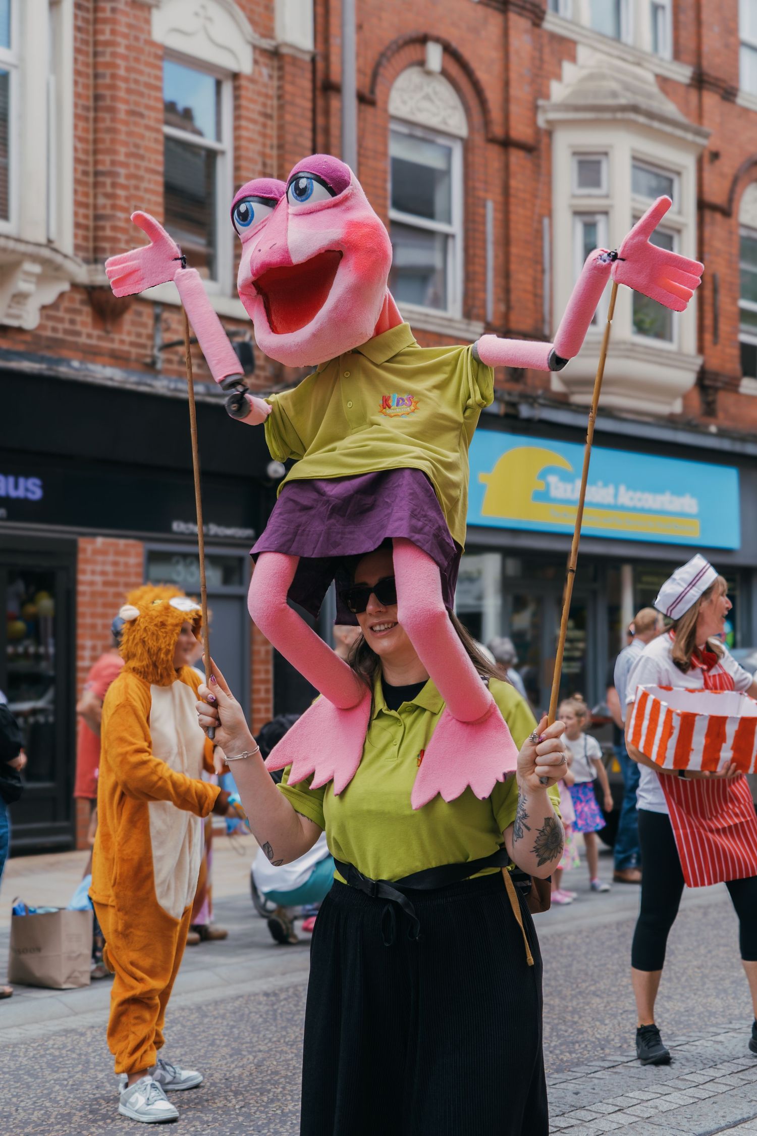 Pink puppet character performing street entertainment in yellow shirt and black pants on a brick-lined city street.