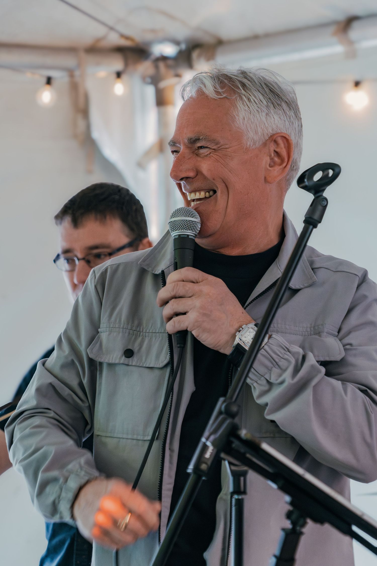Speaker holding microphone and smiling while presenting at outdoor event under string lights.