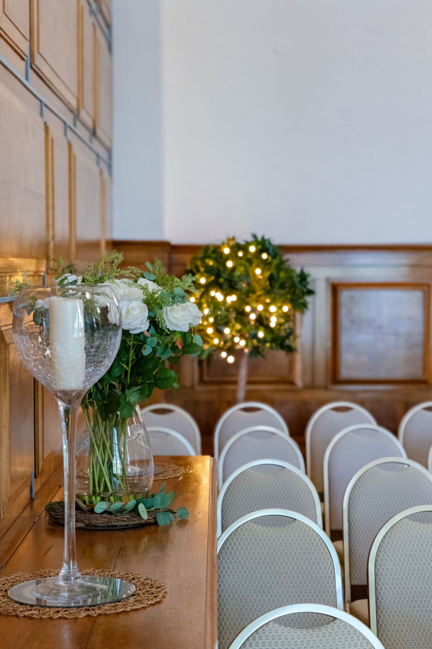 White floral centerpiece in glass vase with lit wreath and chairs arranged for elegant indoor event setup.