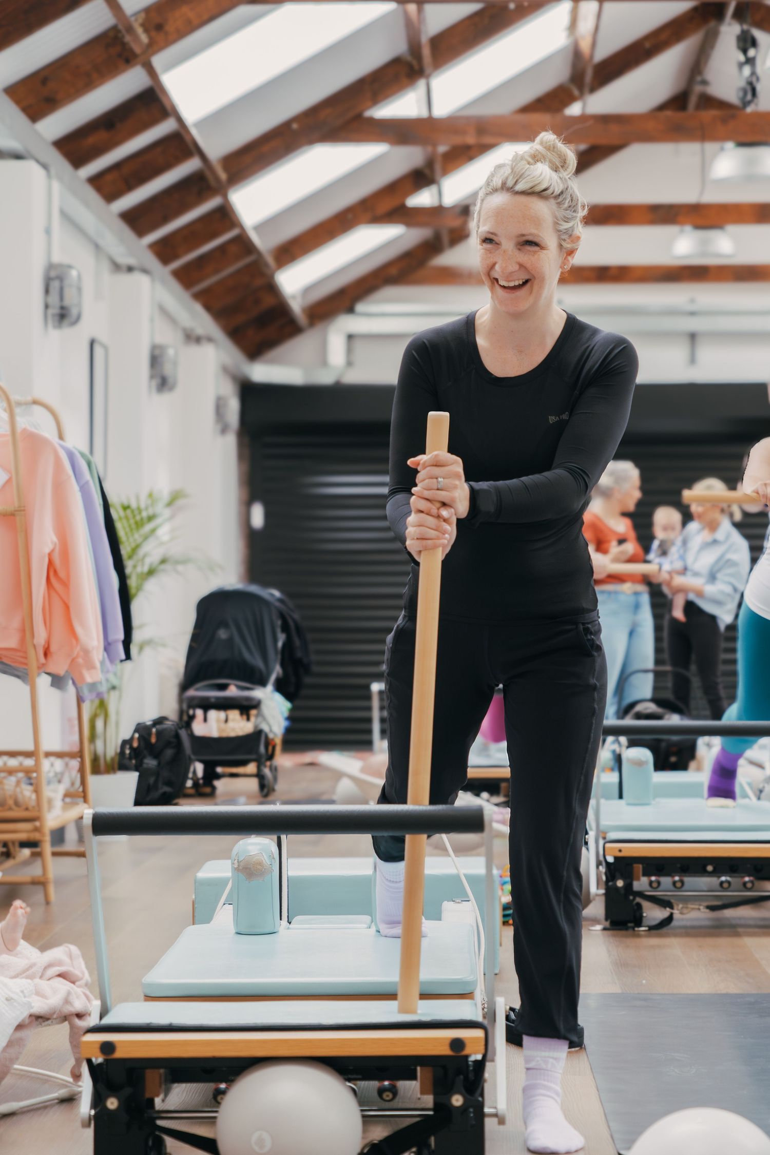 Fitness instructor in black athletic wear demonstrates proper exercise form on Pilates equipment in a bright studio space.