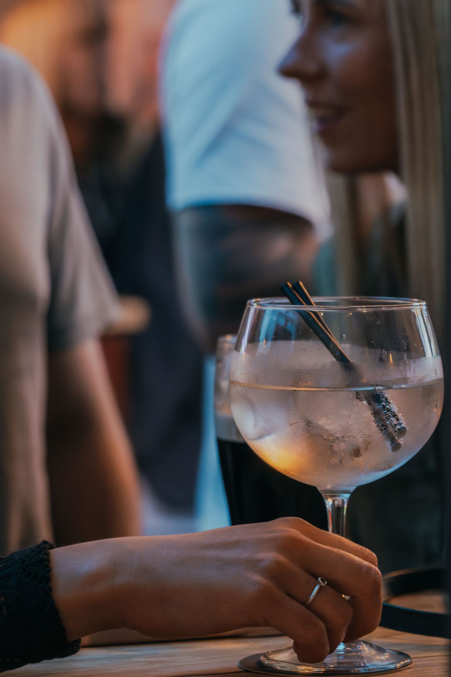 A hand holding a large glass of gin and tonic garnished with botanicals in a dimly lit bar setting.