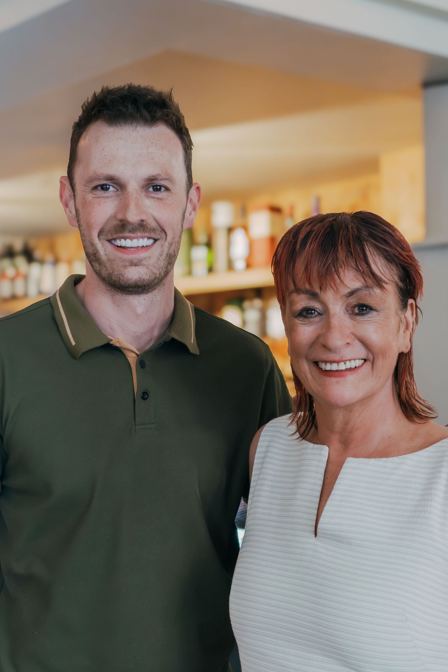 Two people standing together in a restaurant interior with bottles and shelves visible in the background.