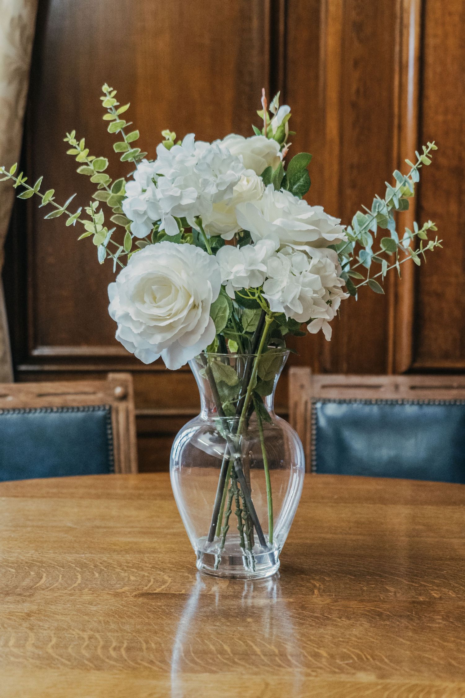 A glass vase with white roses and eucalyptus stems sits on a wooden table with blue chairs in the background.