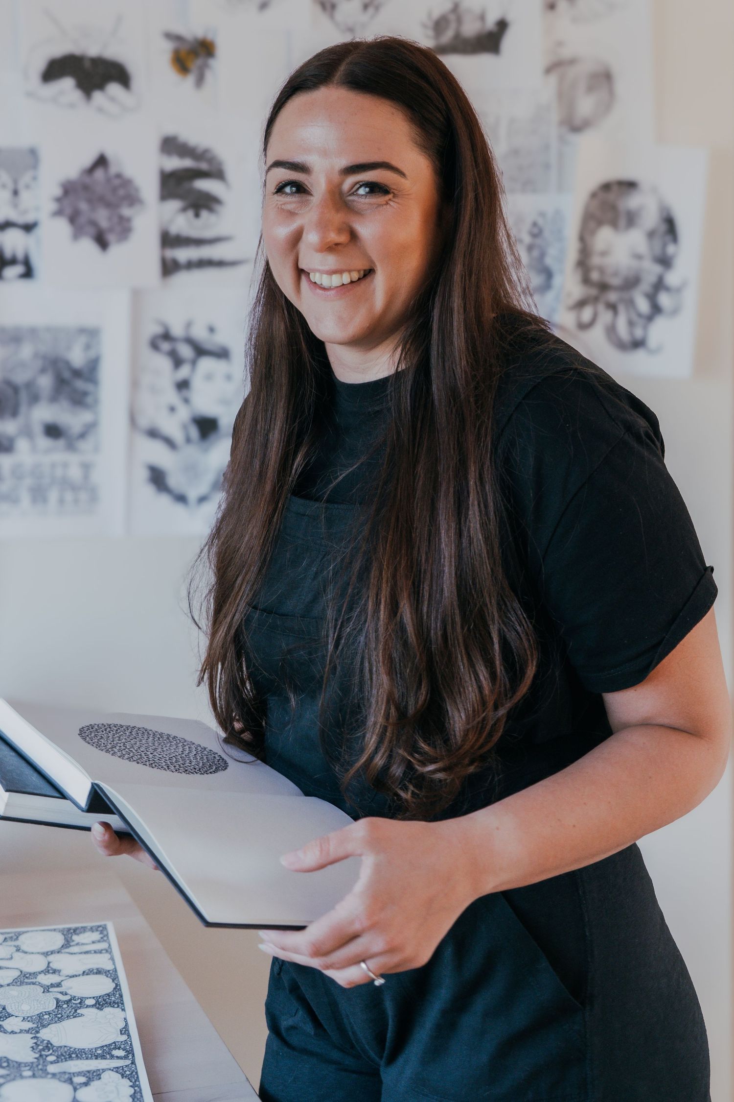 A creative artist stands smiling in a studio workspace with art prints and sketches displayed on the wall behind them.