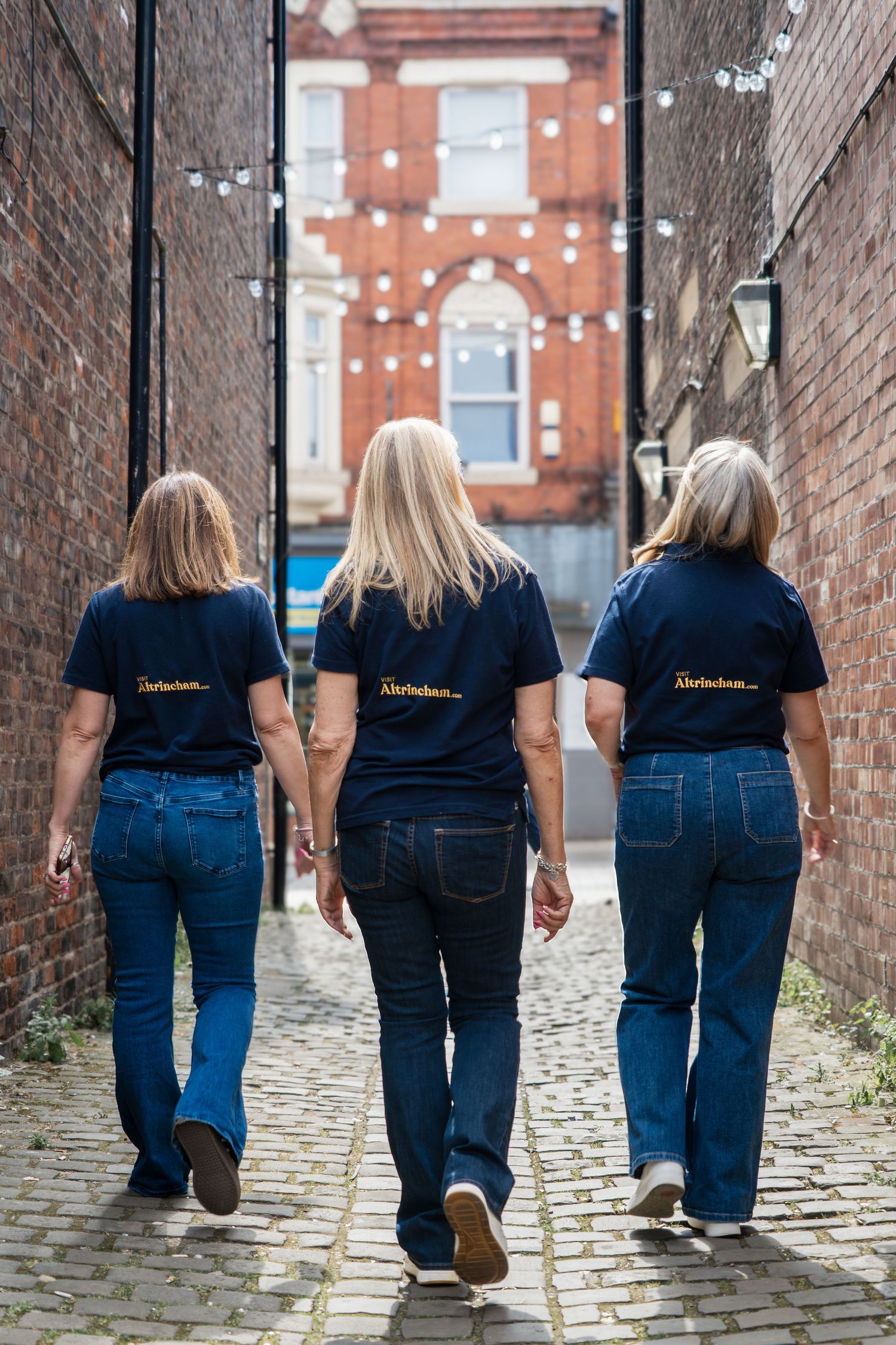 Three people in matching navy t-shirts and jeans walking away down a cobblestone alley between brick buildings.