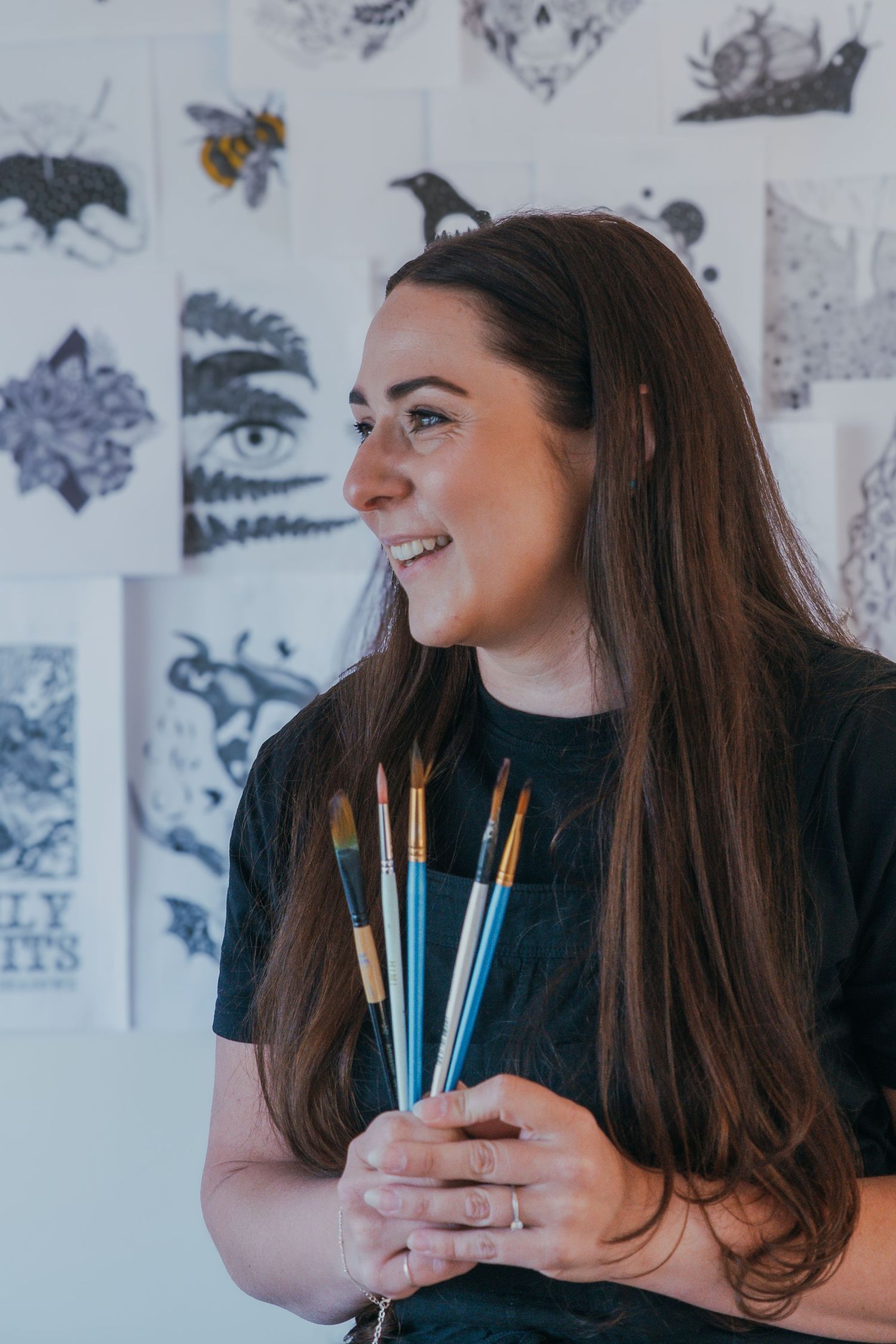 Creative artist holds blue and yellow paintbrushes at art studio with illustrated artworks on the wall behind.