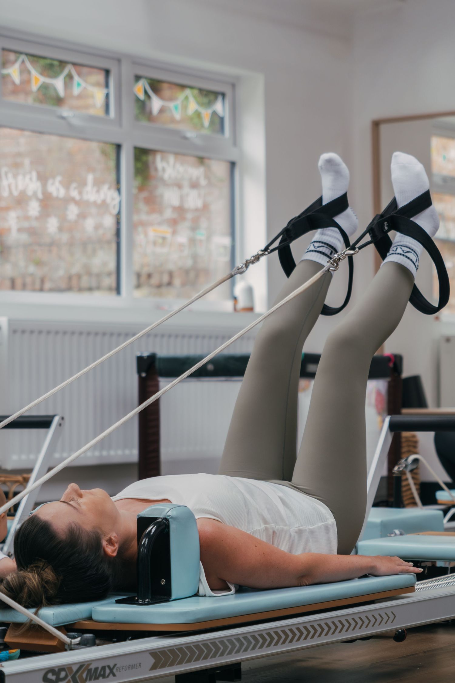 Person exercising on Pilates reformer machine wearing light colored workout clothes in bright studio setting.