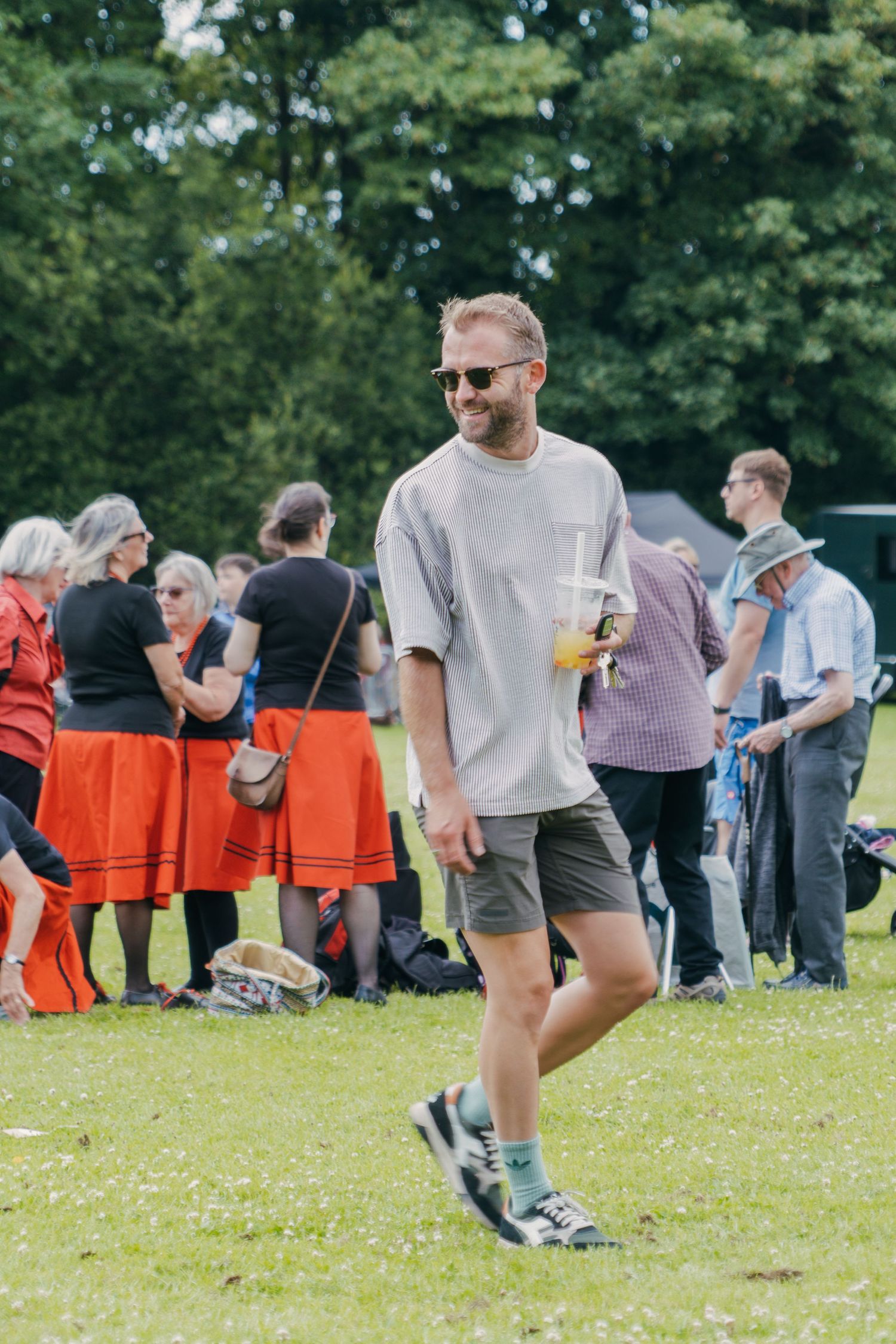 People gathering outdoors at a social event with bright orange chairs scattered on grassy lawn in summer setting.