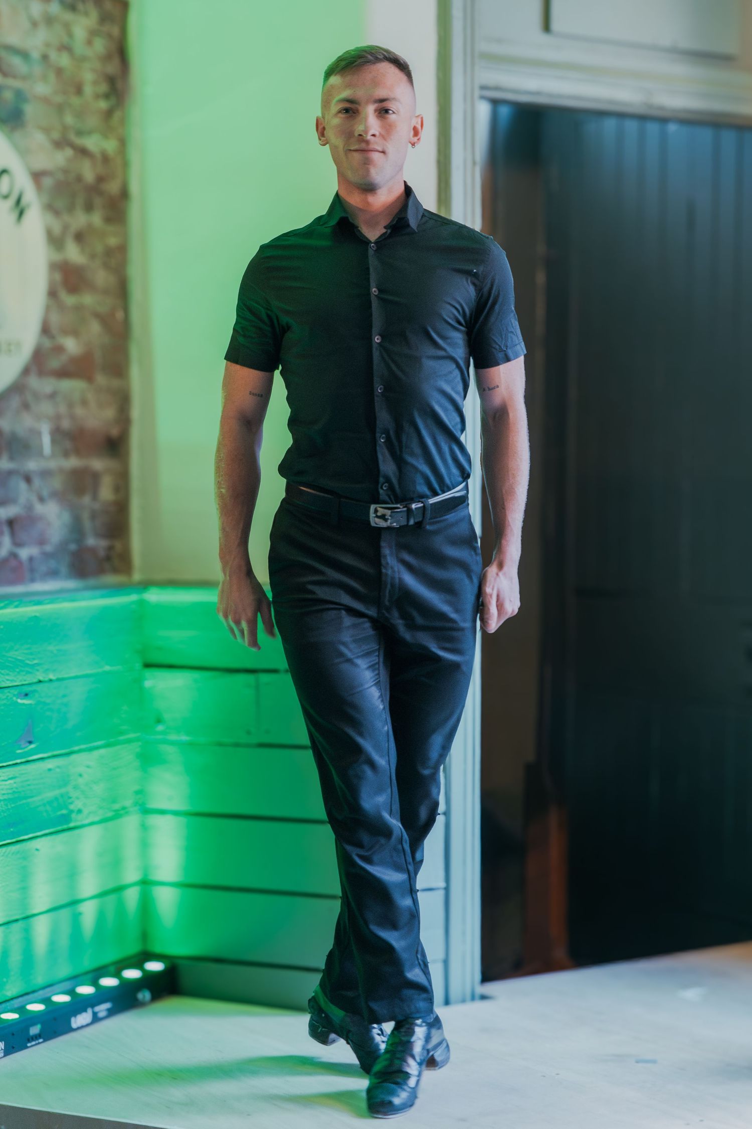 Person wearing all black attire stands confidently against brick wall with green lighting in modern interior space.