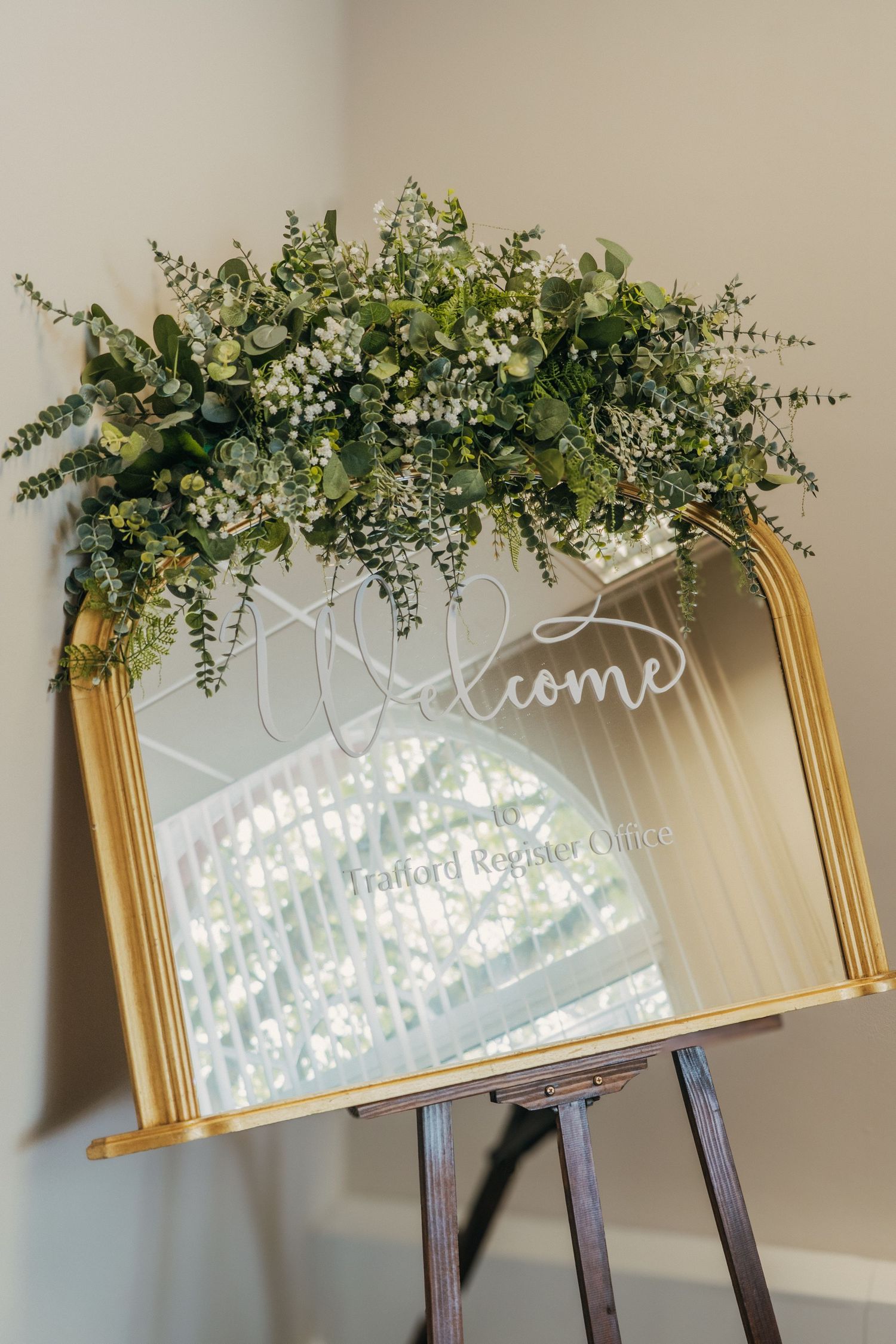 Welcome sign with gold mirror frame decorated with lush greenery and eucalyptus on wooden easel stand.