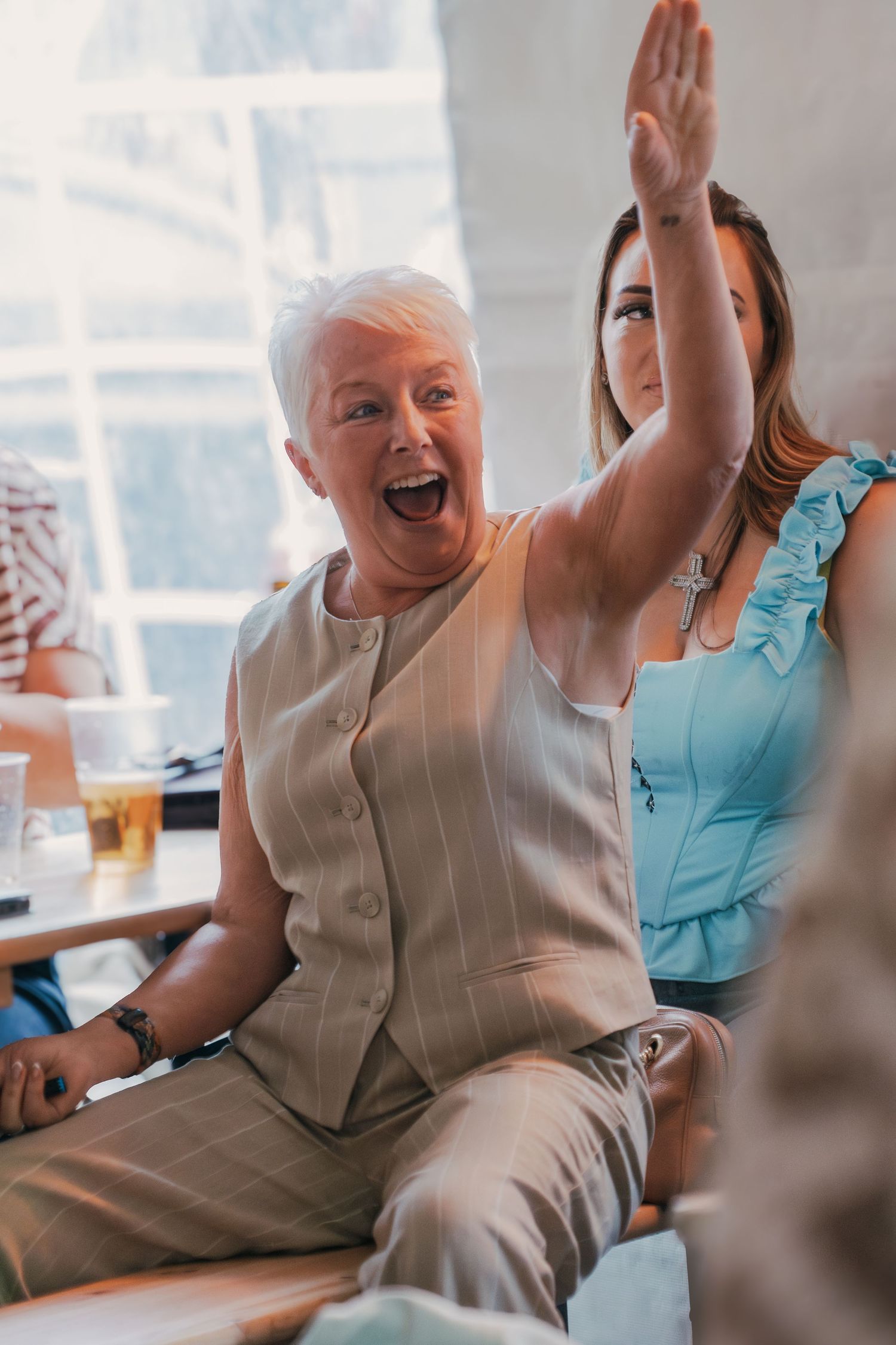 Senior in light clothing cheers enthusiastically while seated during what appears to be a celebratory gathering.