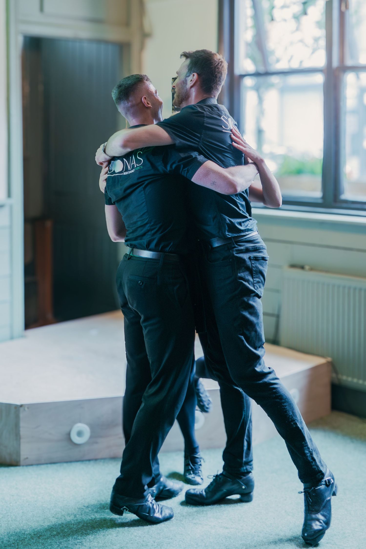 Two dancers in matching dark outfits practice a close embrace dance hold in a naturally lit studio space.