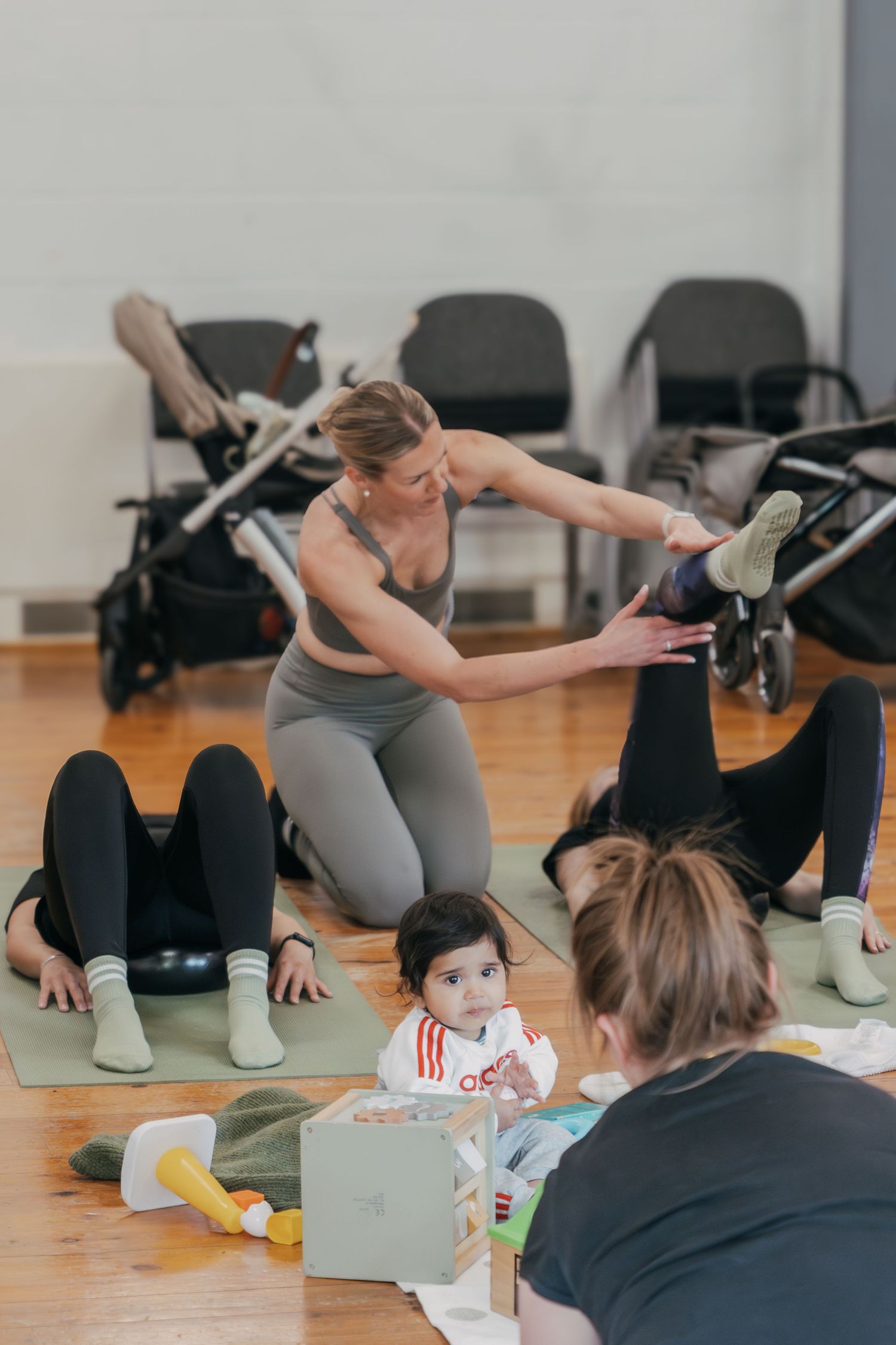 A fitness instructor leads a mom and baby exercise class in a studio with strollers and workout mats on wooden floors.