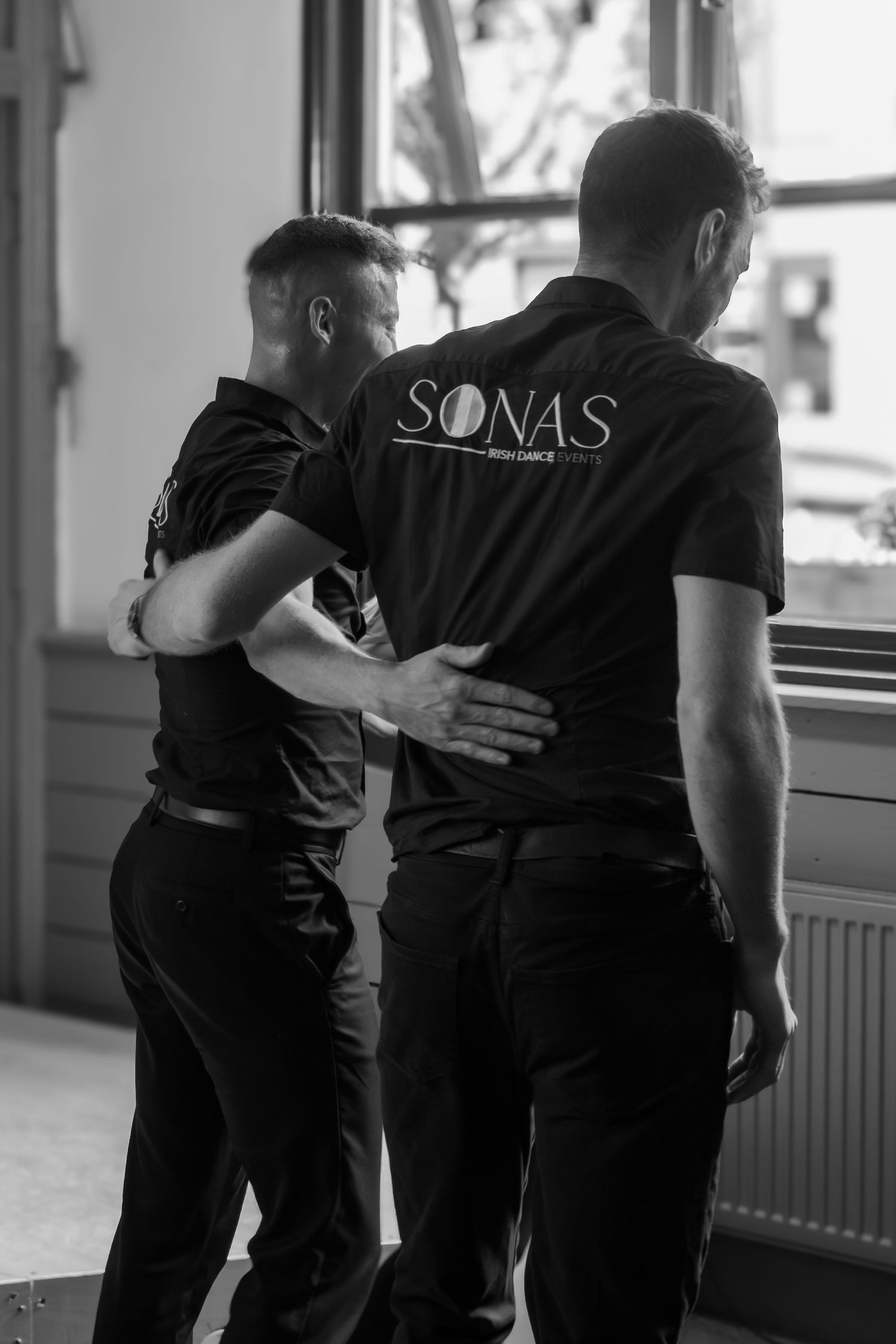 Two people in matching black SONAS t-shirts embrace while dancing in a studio with window light behind them.