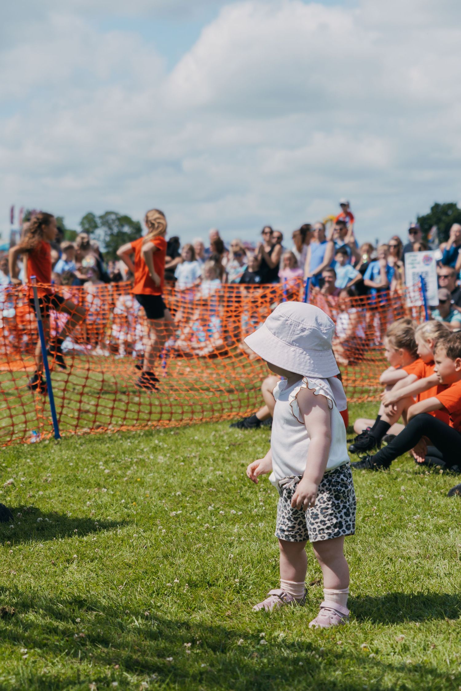Kids play and run on green grass field during outdoor event with orange safety barrier visible in background.