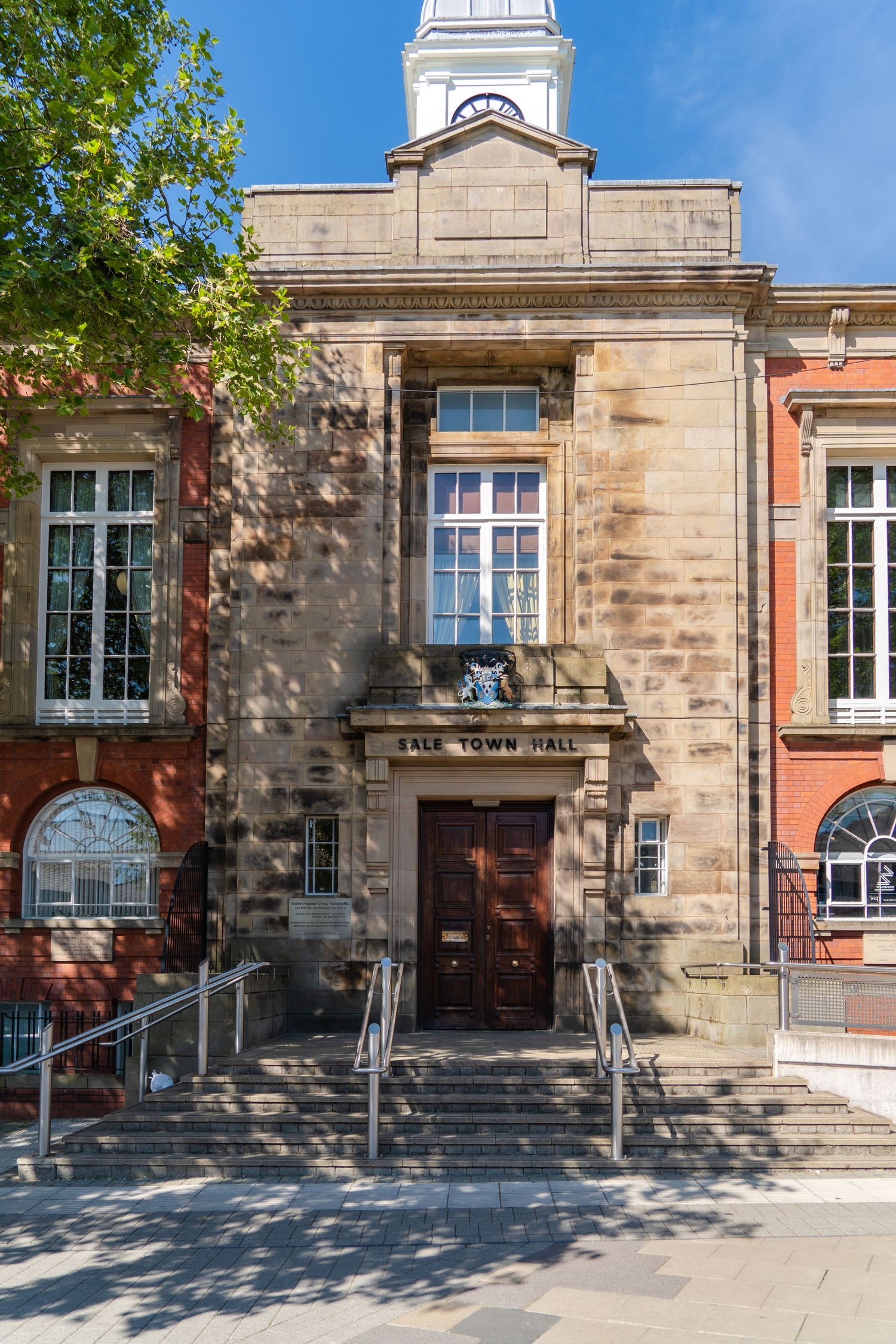Historic stone building entrance with white cupola, arched windows, and central wooden door with handrails leading to steps.