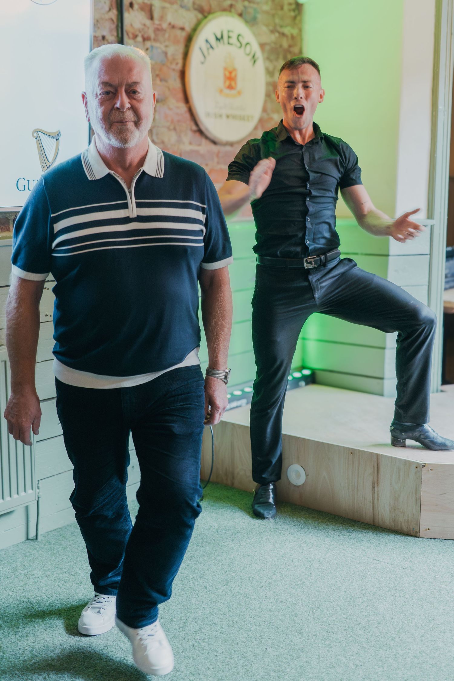 Two men in casual attire pose indoors with contrasting expressions - one calm and one energetically jumping.