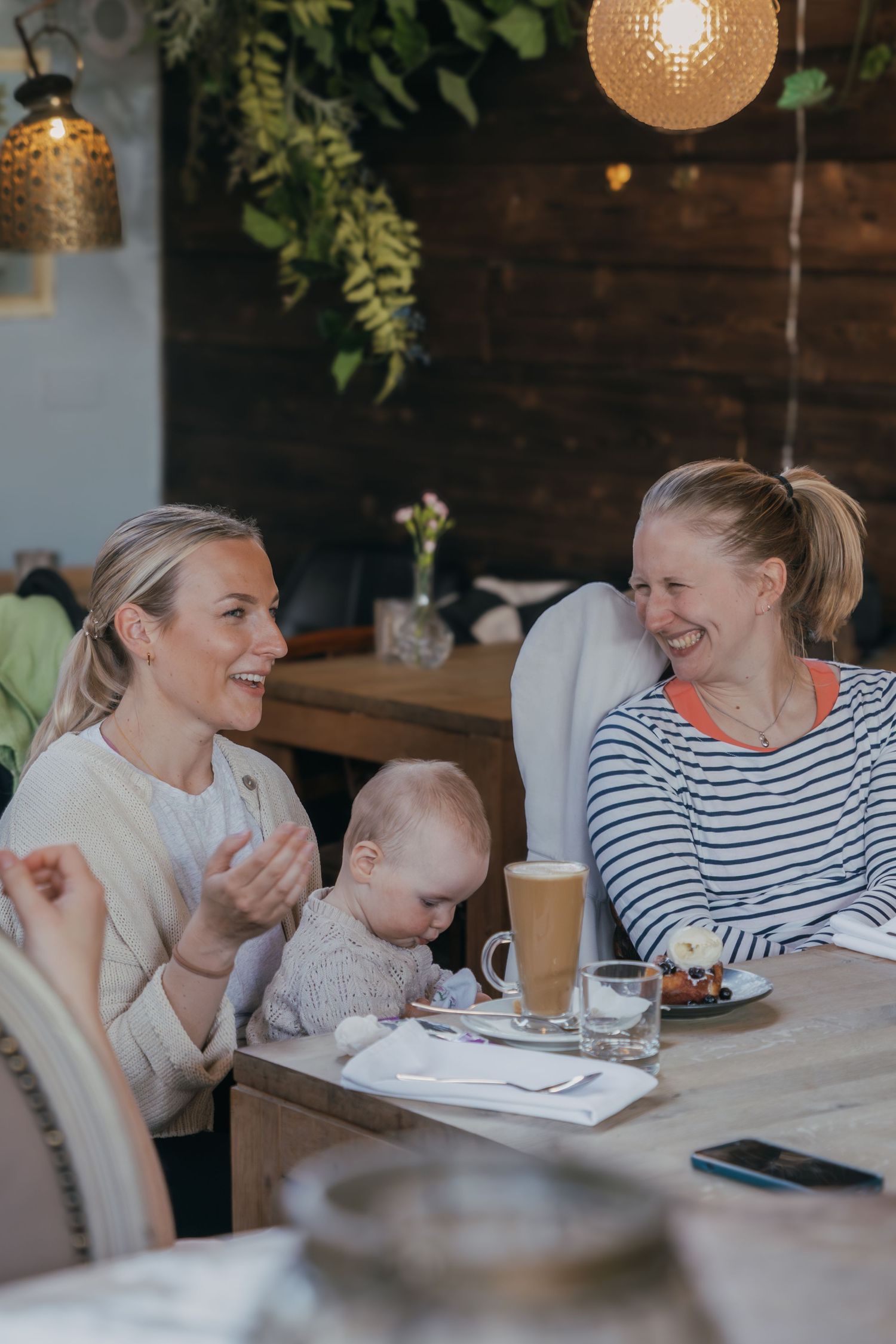 Three friends enjoying coffee and conversation at a cozy cafe with exposed brick walls and hanging pendant lights.
