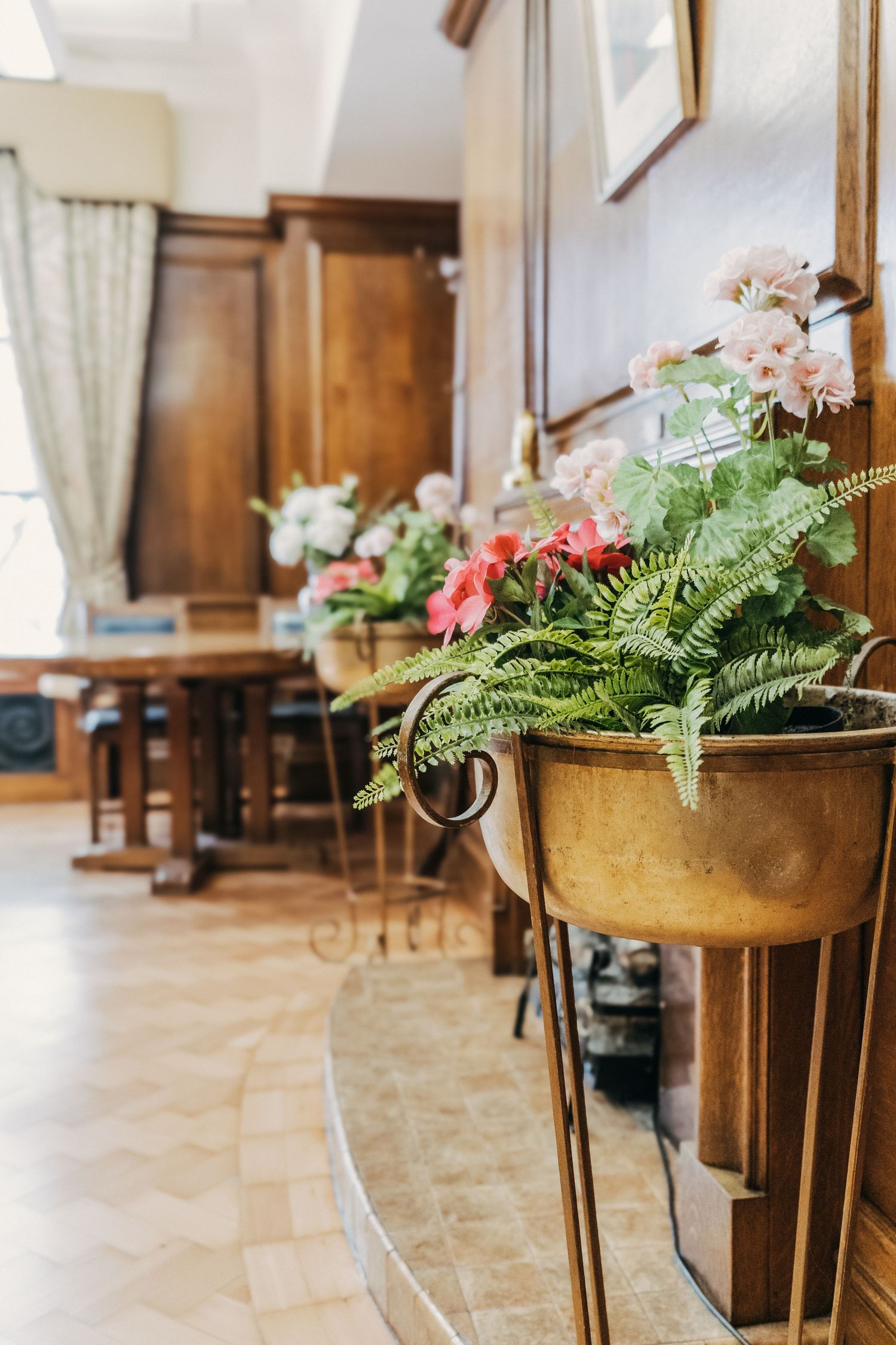 Potted ferns and pink flowers in brass planters line an elegant hallway with wooden trim and tiled flooring.