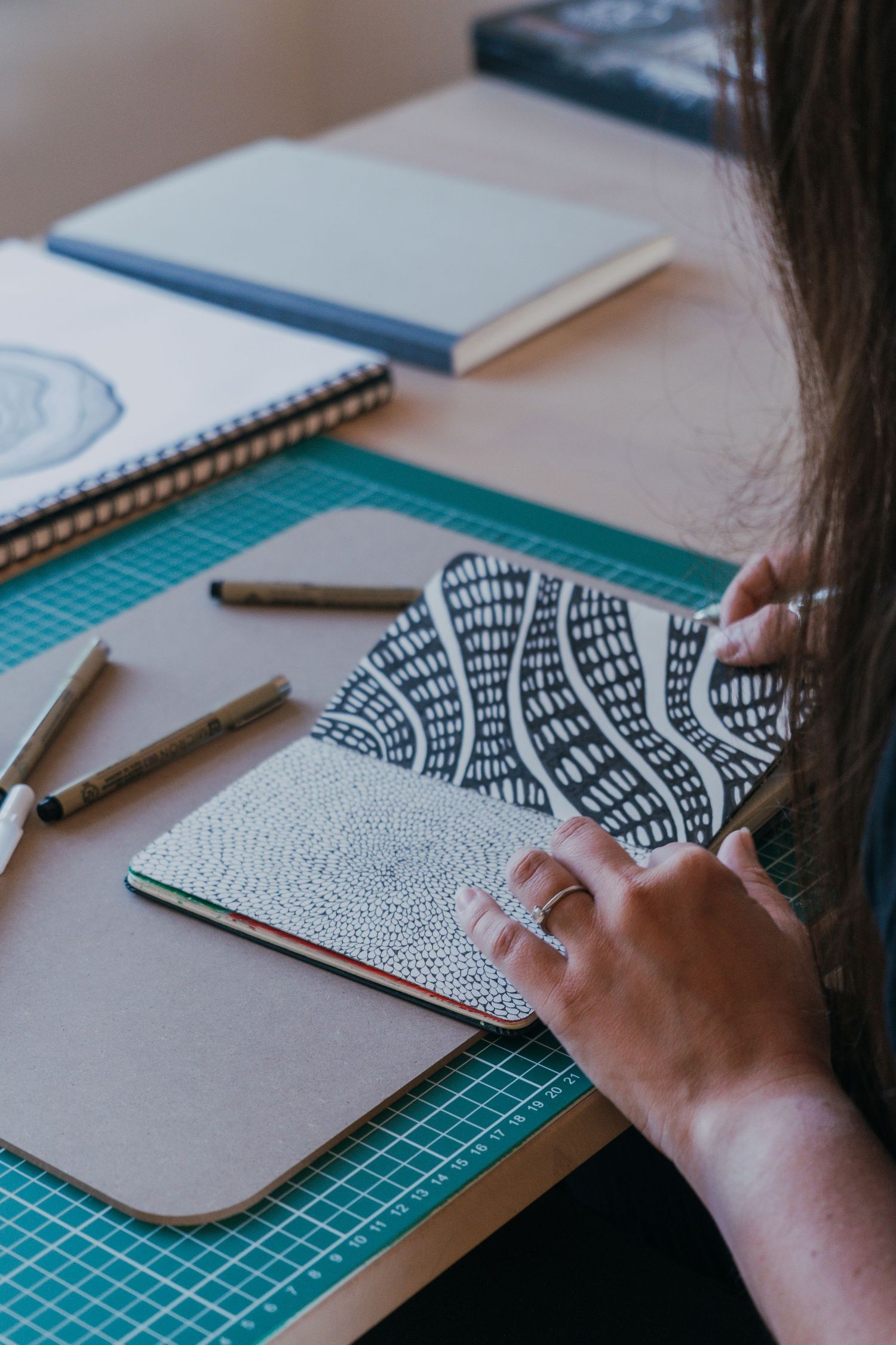 Artist drawing intricate black and white pattern in sketchbook on green cutting mat with art supplies nearby.