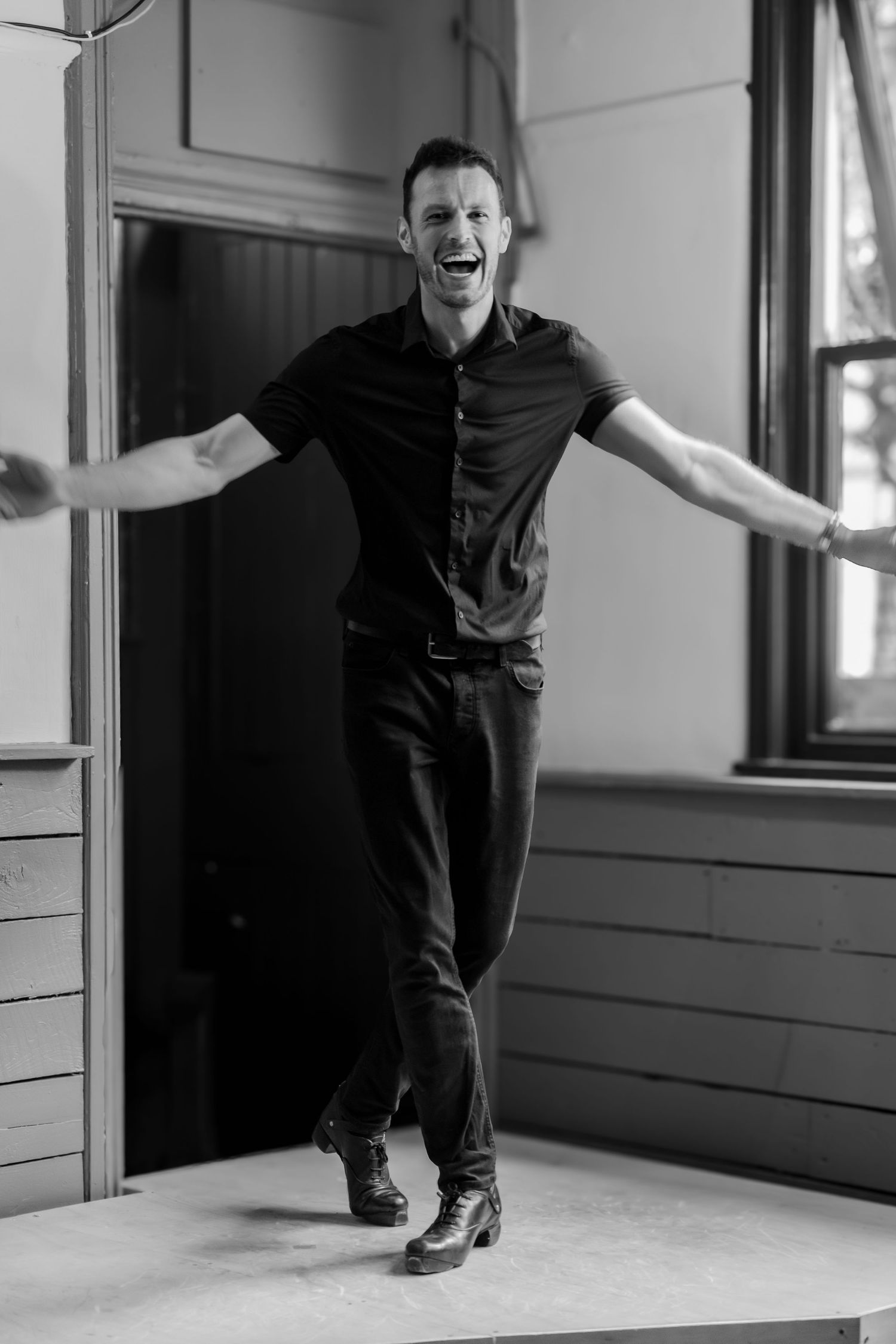 Black and white photo of a person jumping joyfully in a sunlit room wearing dark clothing.