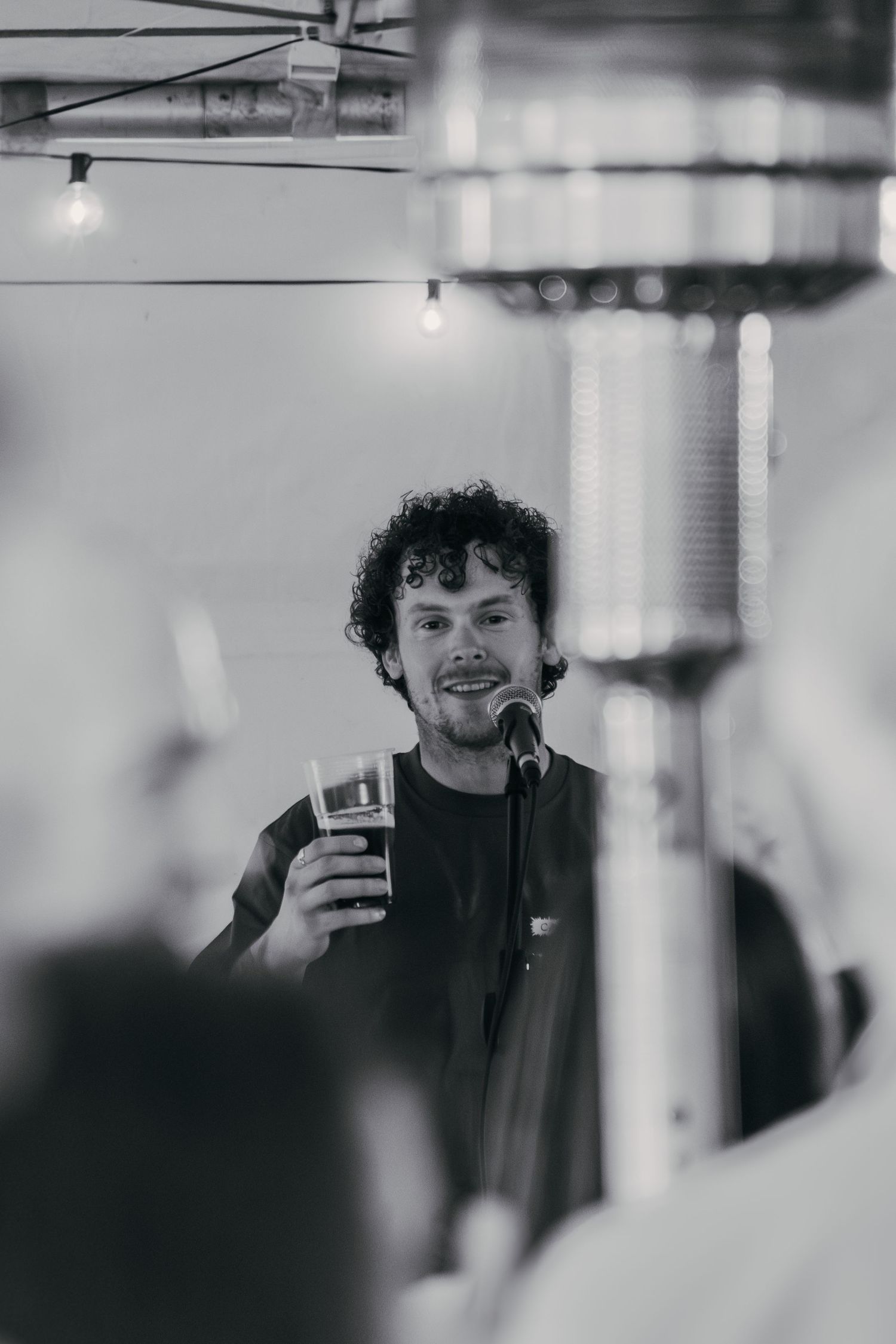 A performer holds a beverage while speaking into a microphone in a black and white photograph.