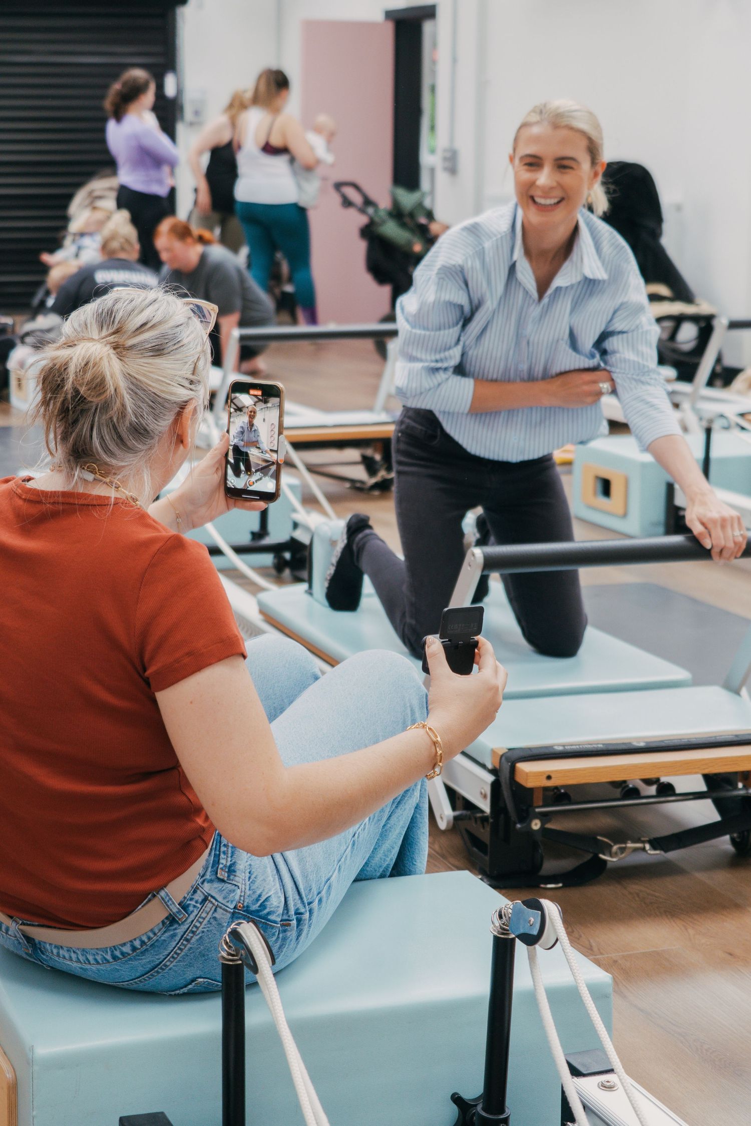 Two people using exercise equipment in a bright fitness studio with pilates reformer machines in the background.