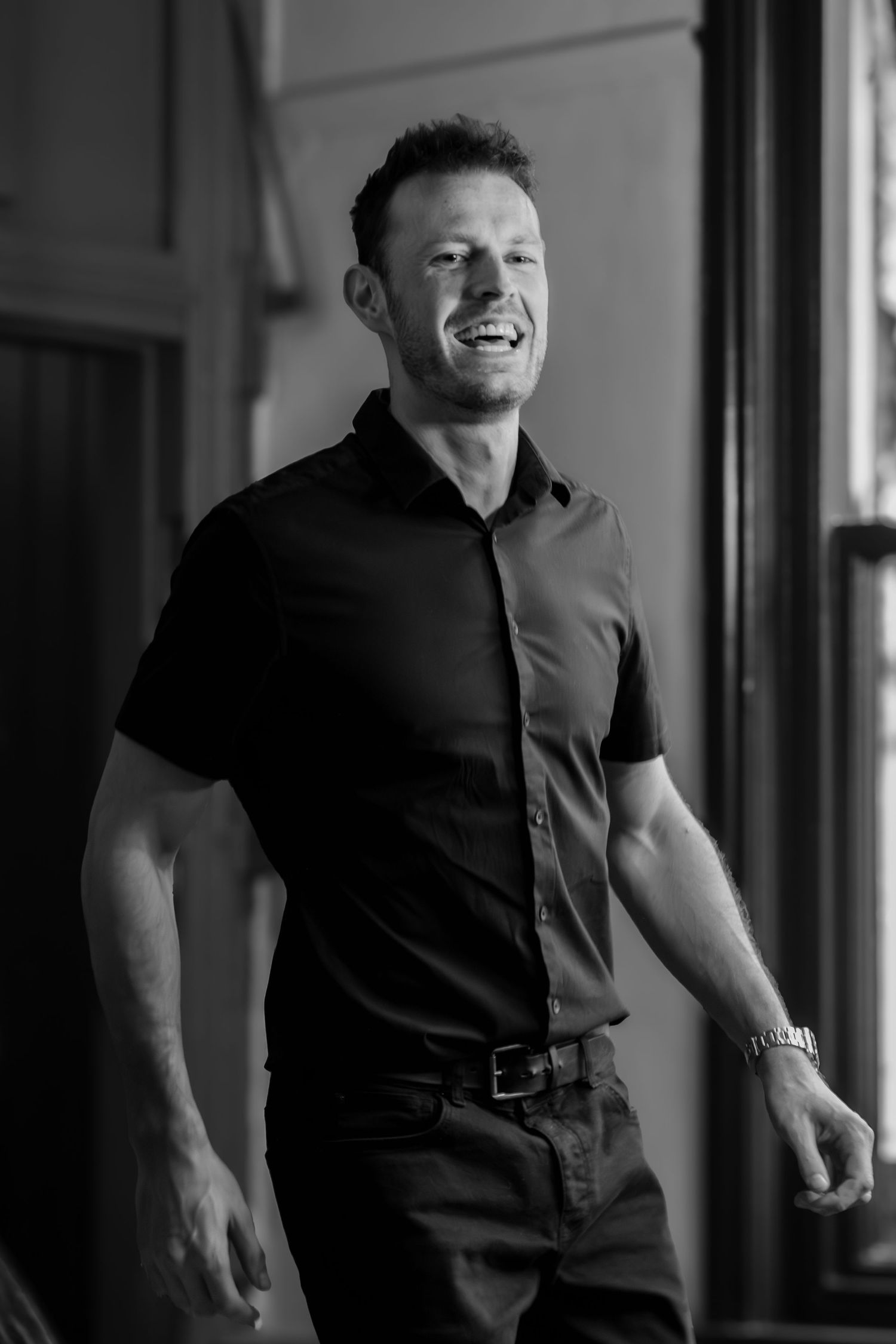 Black and white portrait of smiling man in dark short-sleeve button-down shirt standing by window.
