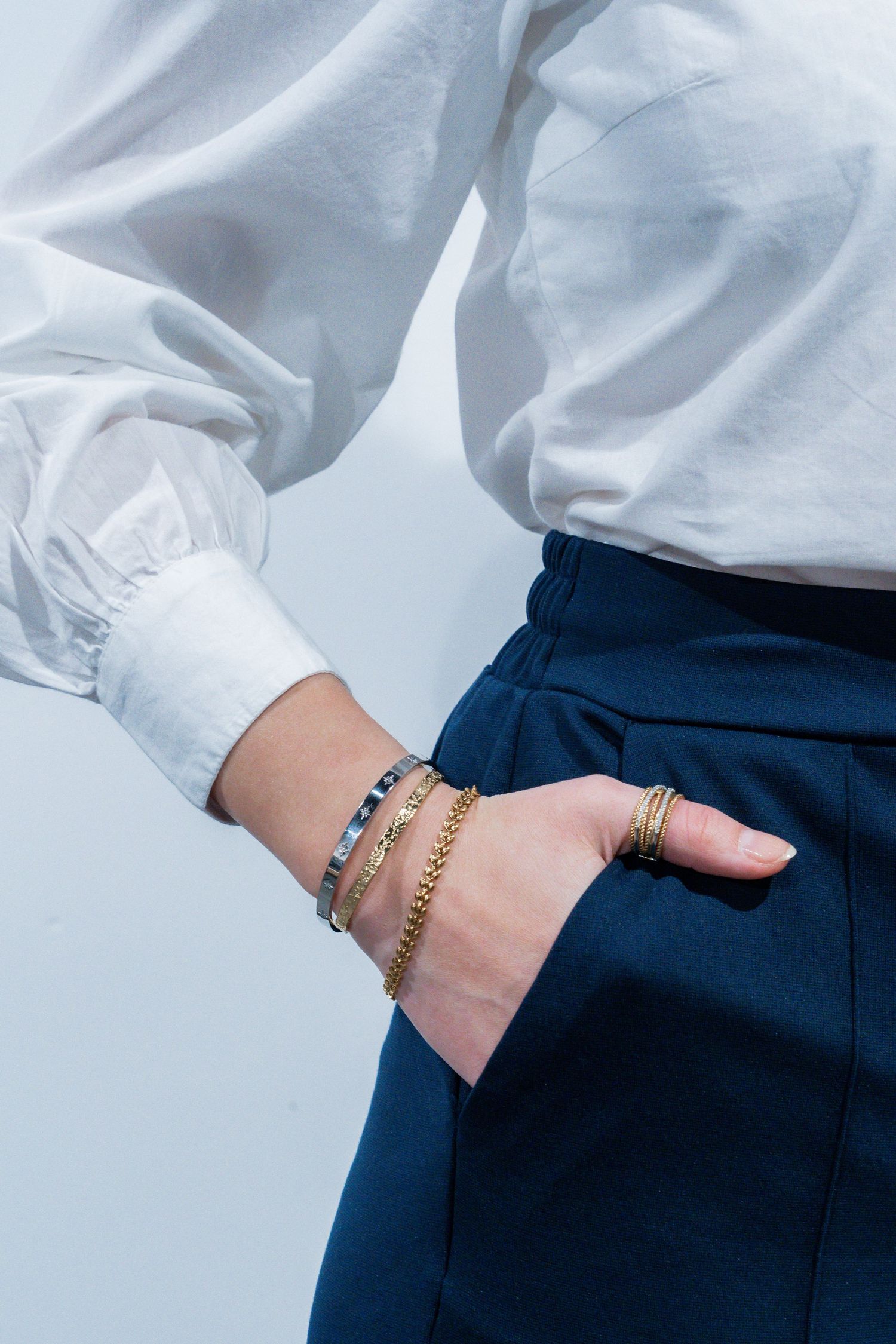 A close-up of delicate gold bracelets and rings styled with a white shirt and navy blue pants.