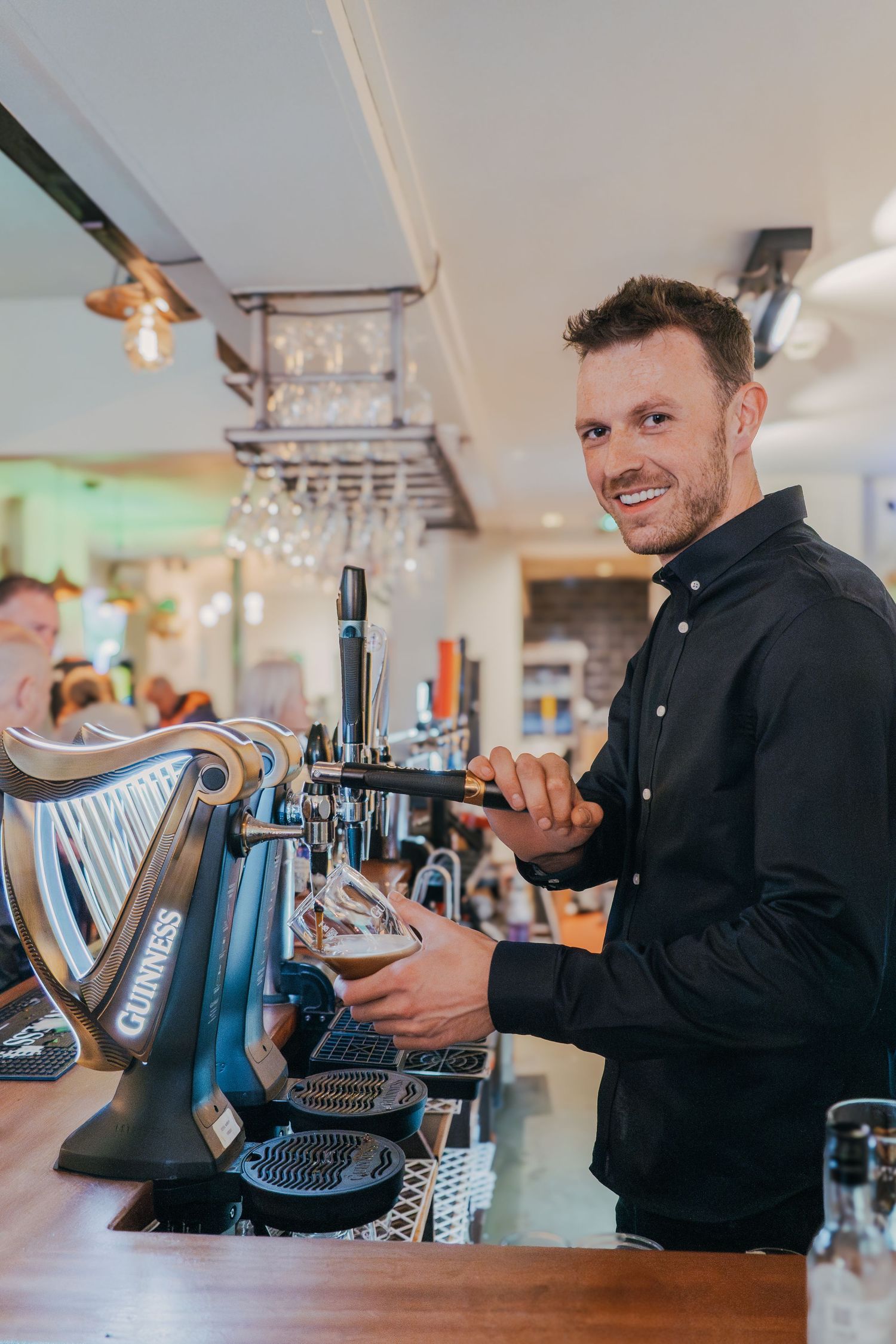 A bartender in a black shirt pours a draft beer from a tap at a modern bar with overhead glass storage.