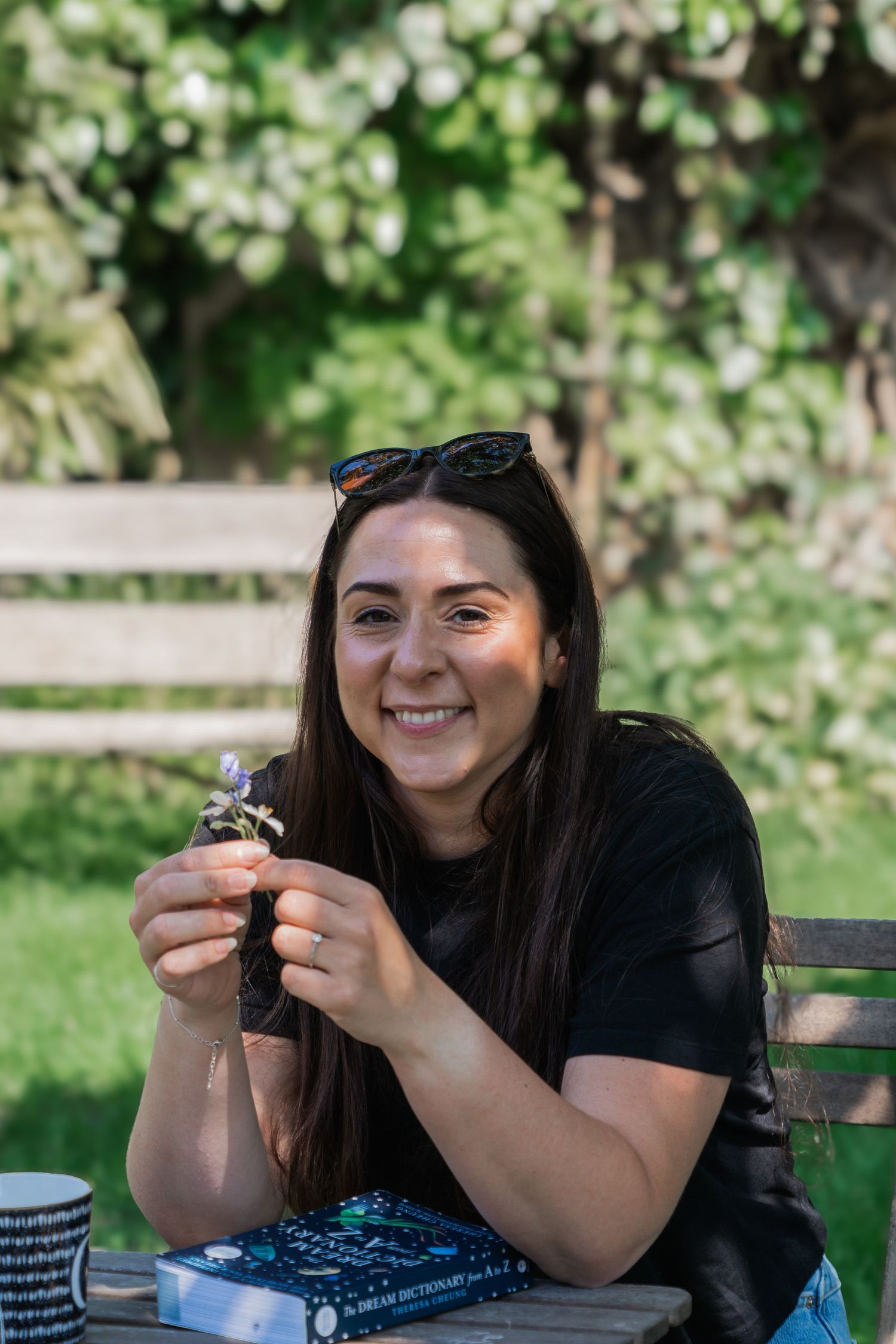 Person in black t-shirt sits outdoors at table smiling while holding something small in their hands on sunny day.