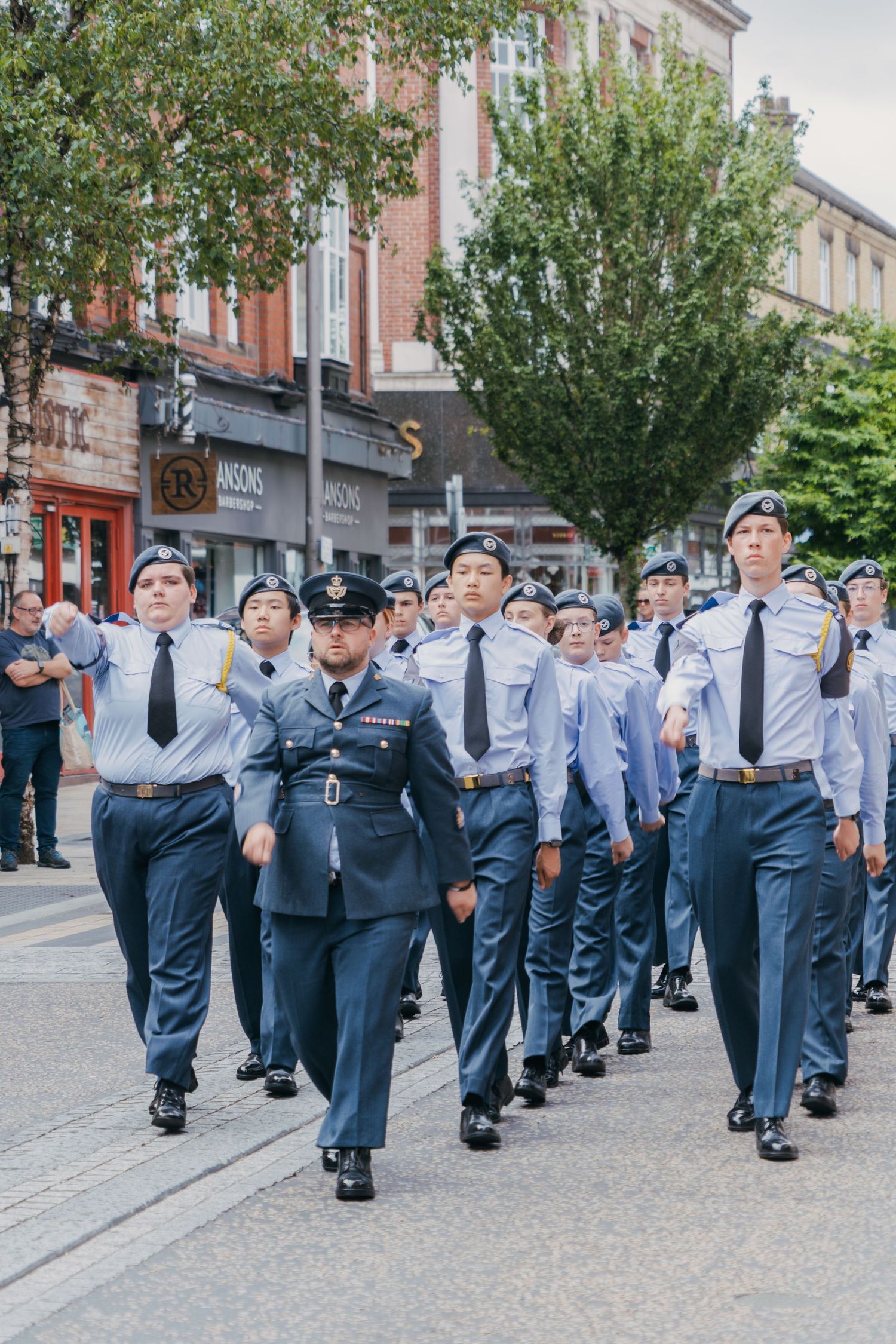 A group of uniformed cadets marching in formation down a city street lined with trees and buildings.