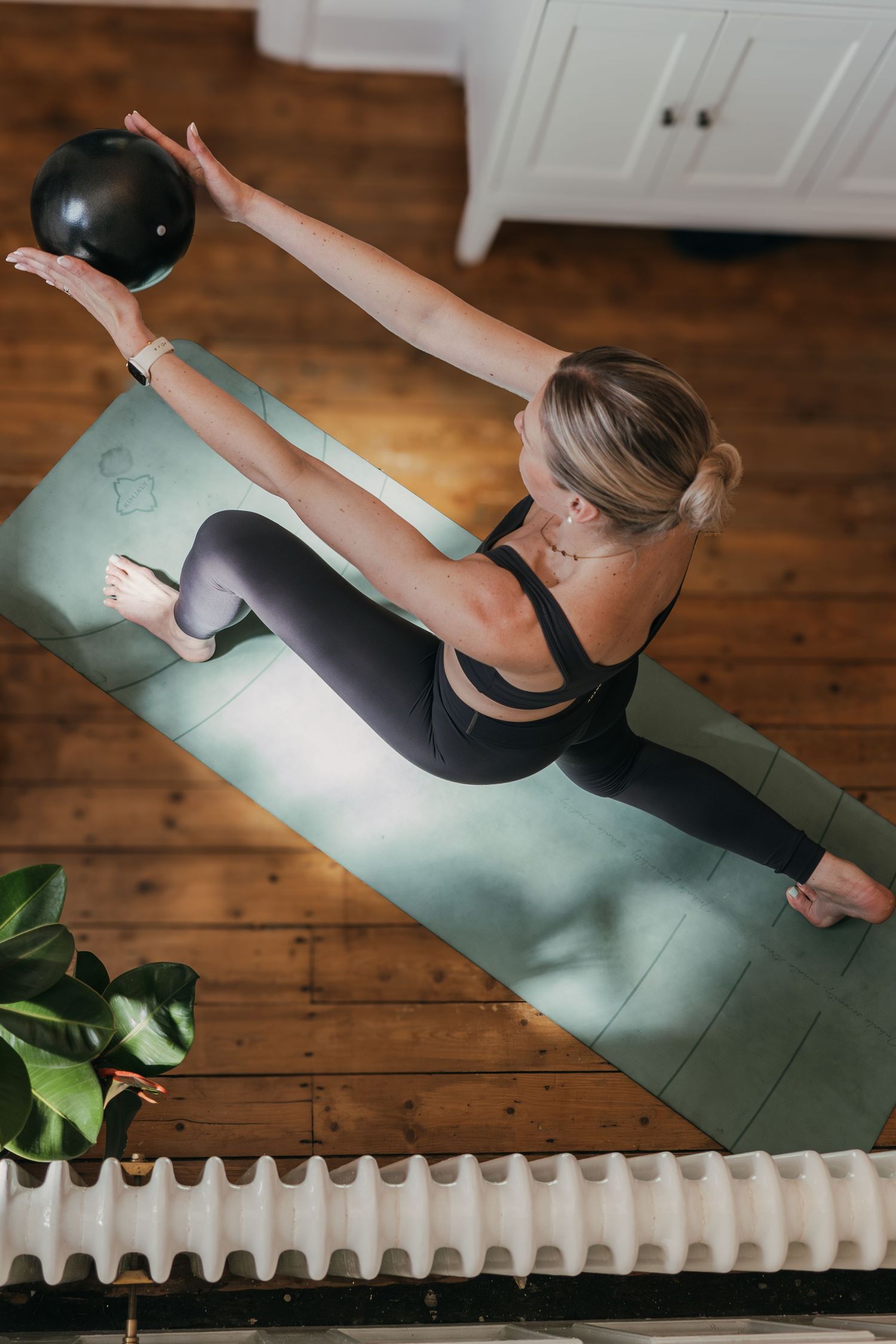 Overhead view of person exercising with medicine ball on yoga mat next to radiator and potted plant on wooden floor.